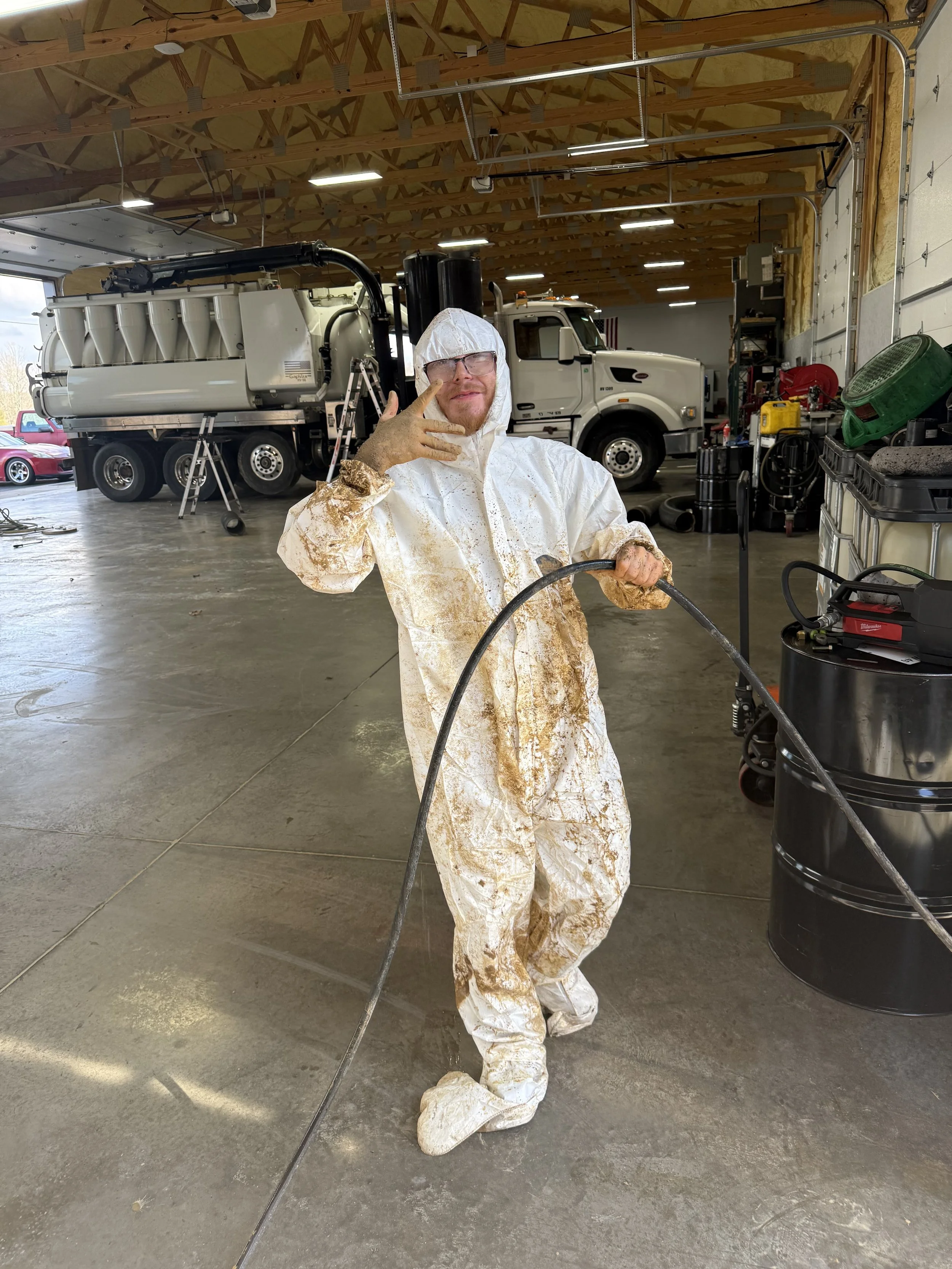 Ethan wearing a tyvek suit. The suit has mud or dirt stains. In the background, there are trucks and industrial equipment.