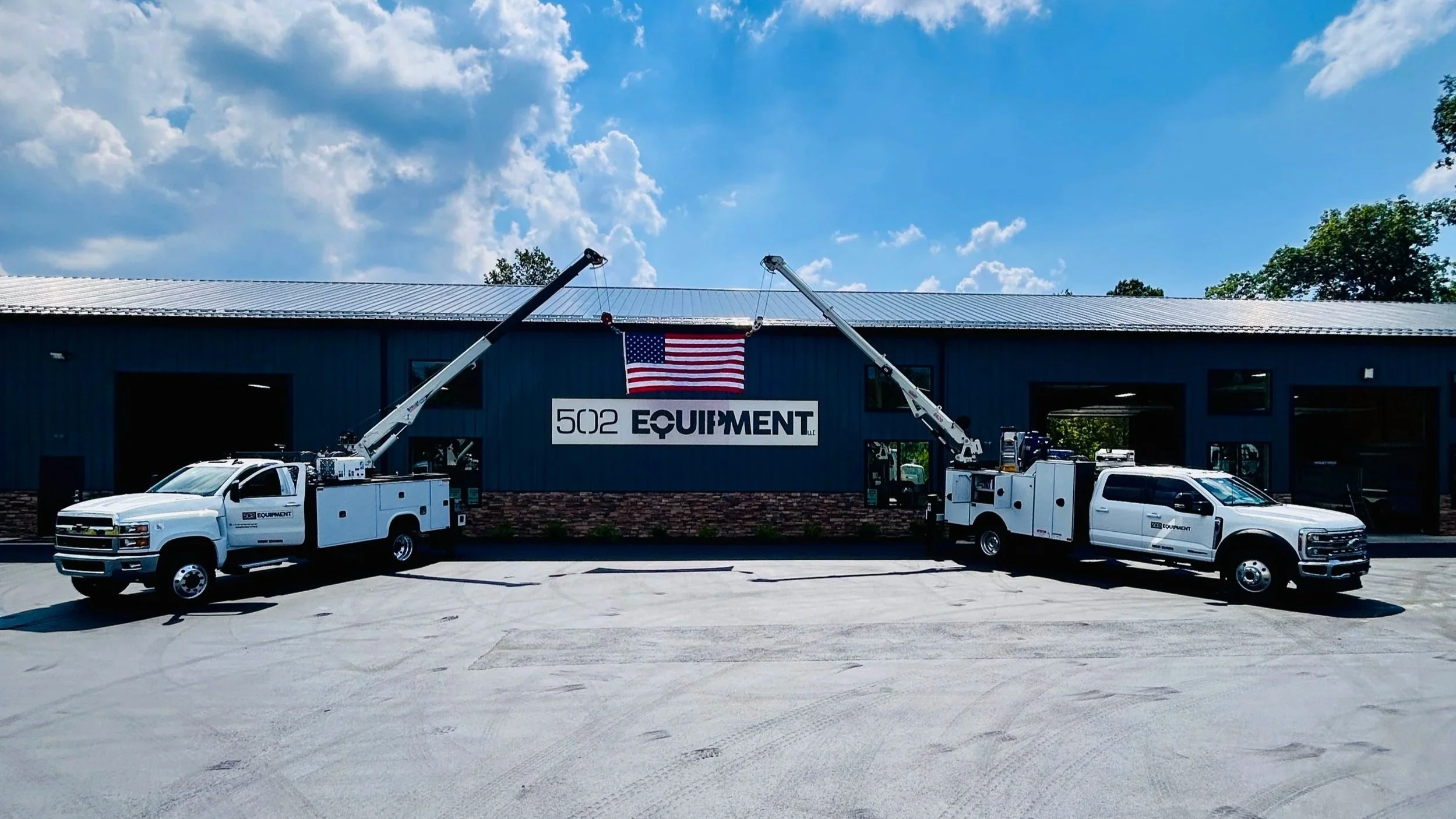 Two white mobile service trucks with cranes extended holding an american flag above the 502 Equipment vac truck service center in LaGrange, KY.