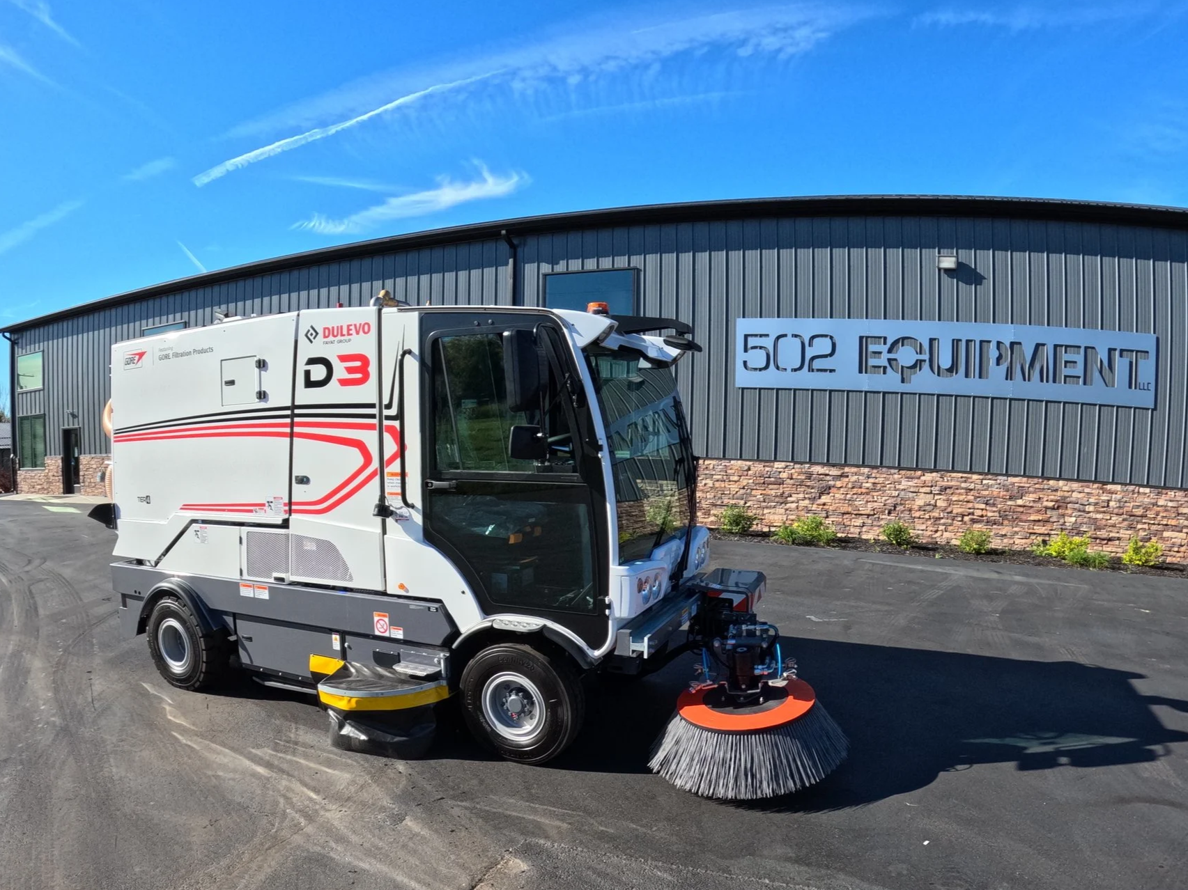 Street sweeping machine in front of a commercial building with a sign reading '502 Equipment LLC' under a blue sky with clouds.