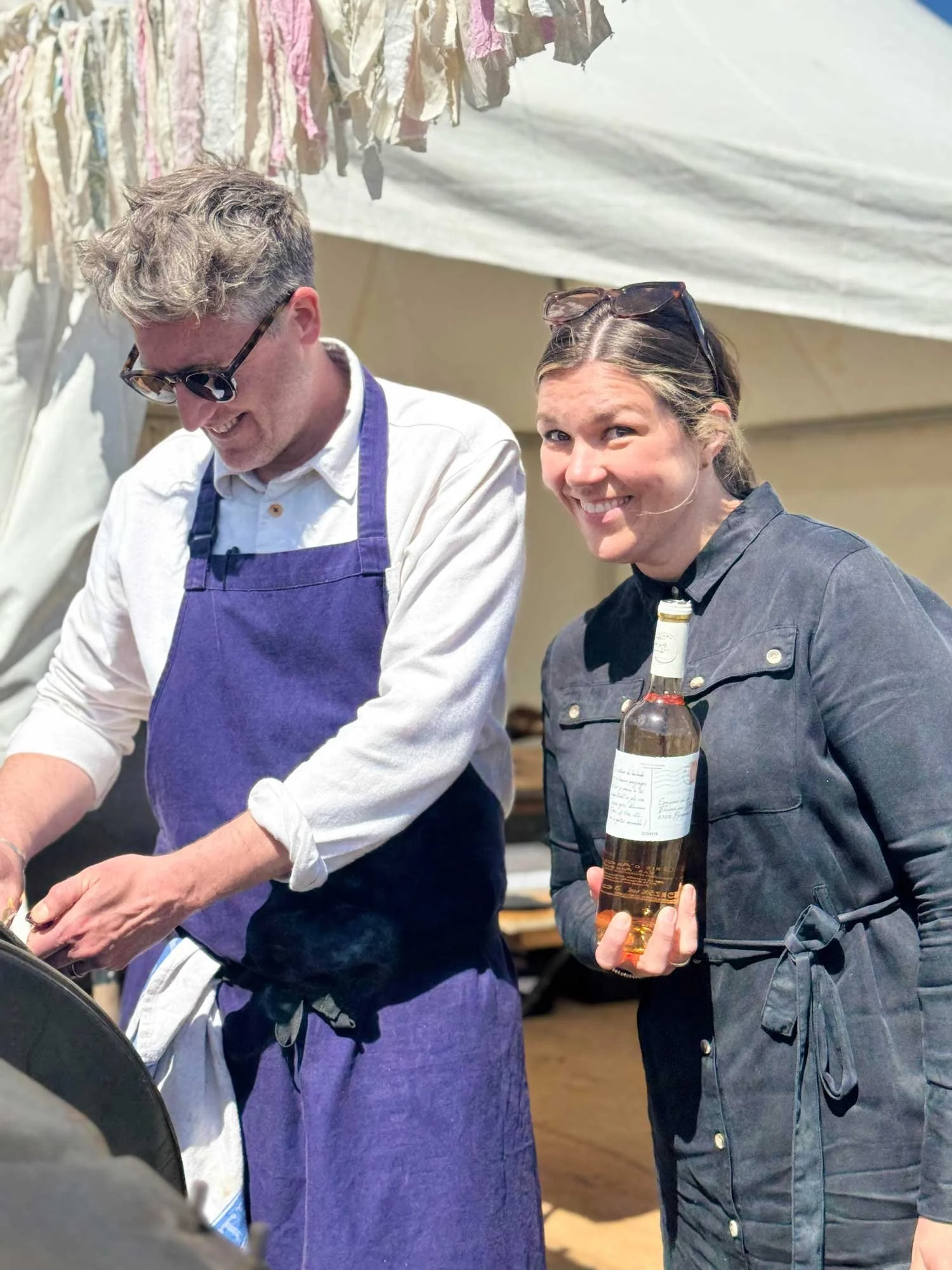 A woman with sunglasses on her head, holding a bottle of wine and smiling, standing next to a man in sunglasses and a purple apron. They appear to be at an outdoor event or market.