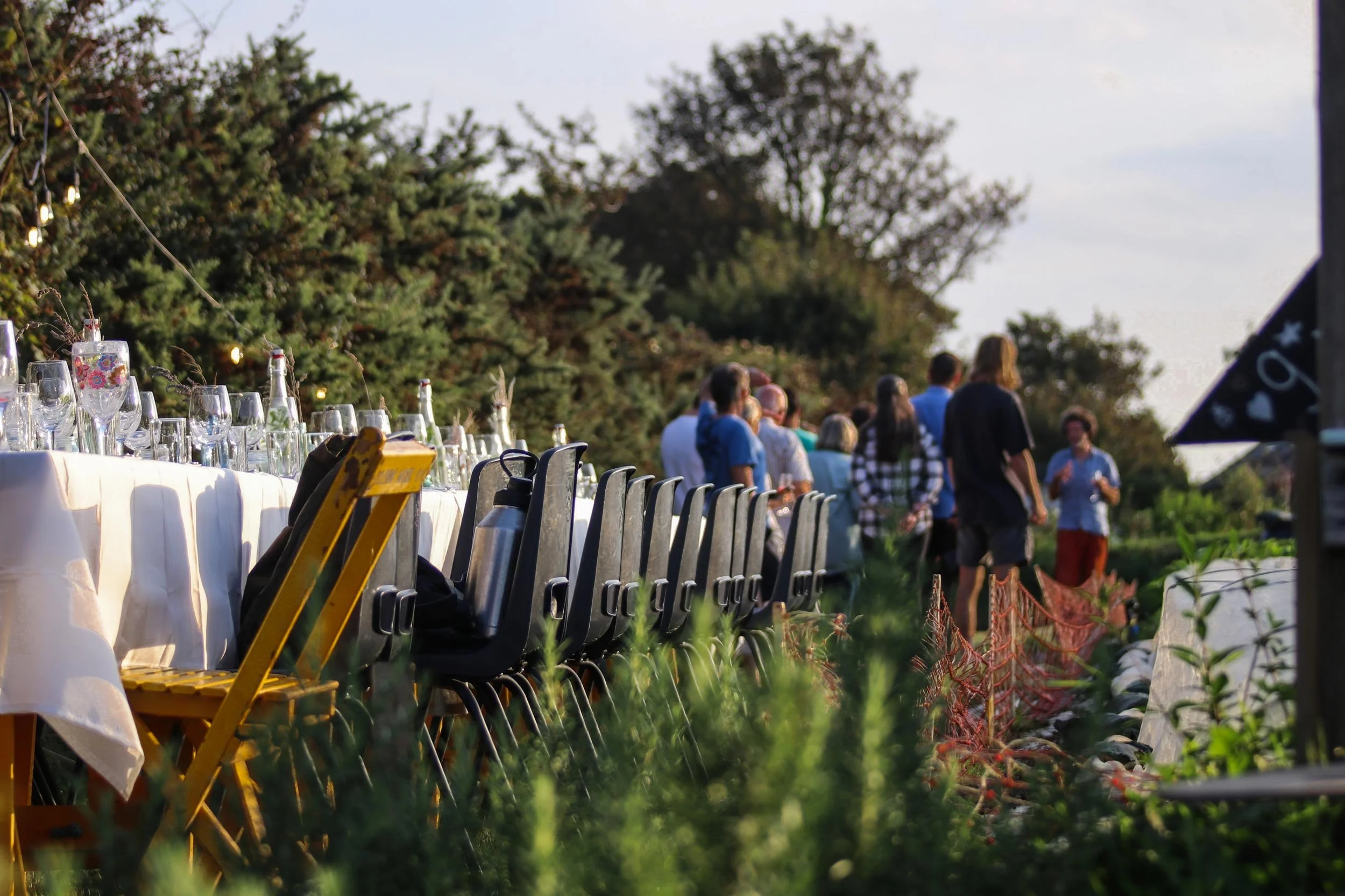 Outdoor event with a long table set with glassware and drinks, surrounded by chairs, with several people standing and talking in the background, near trees and a partly cloudy sky.