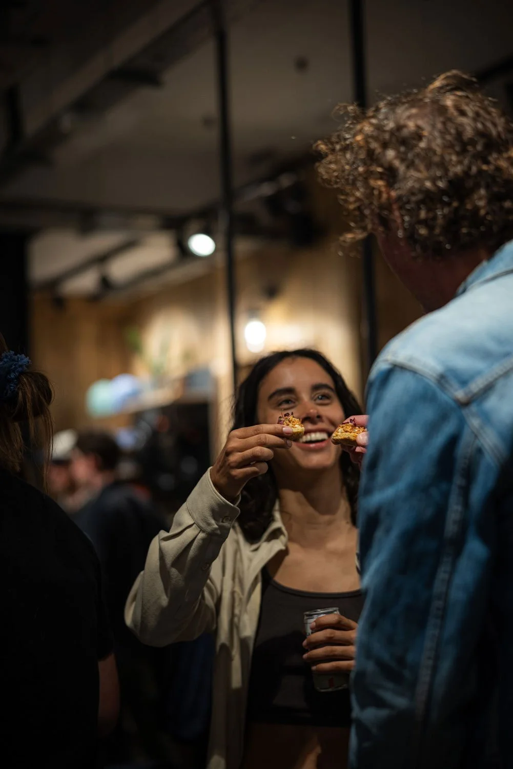 People socializing in a dimly lit bar, with a woman holding a small piece of food towards a man, smiling and engaging in conversation.