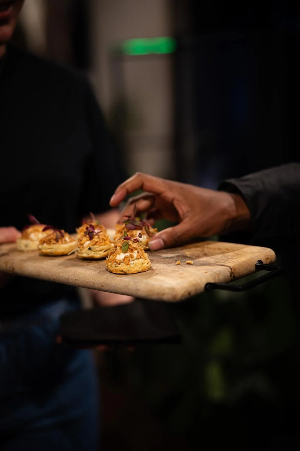 Person's hand placing small appetizers or canapés on a wooden serving board with other people nearby, in a dimly lit setting.