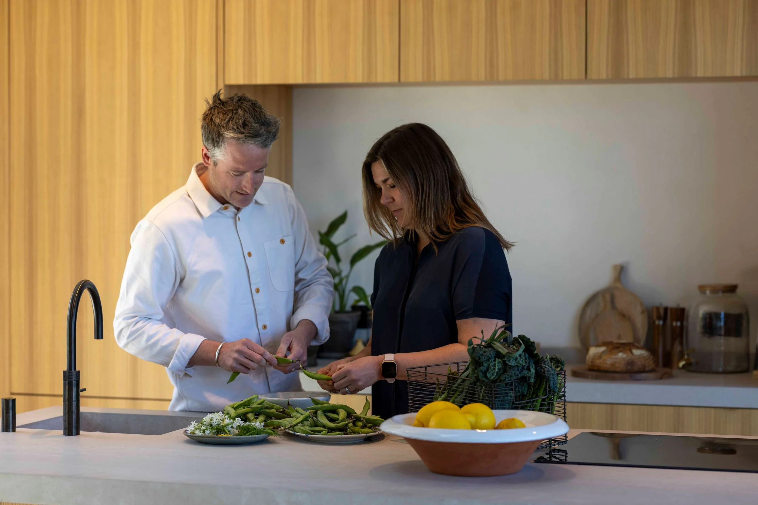 A man and woman standing at a kitchen counter, examining a green vegetable, with various vegetables and flowers on the counter.