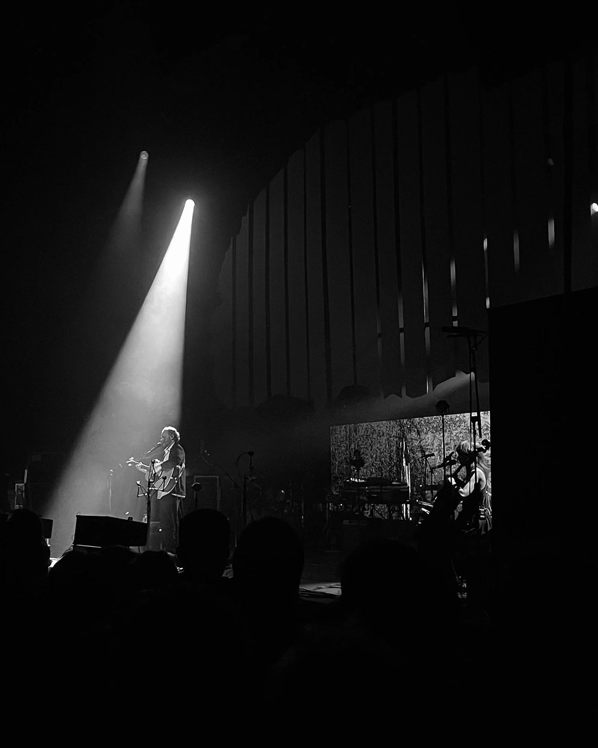 Black and white photo of a musical performance on stage, with a singer playing guitar and singing into a microphone, and other musicians seated with instruments, with vertical curtain in the background and audience in the foreground.