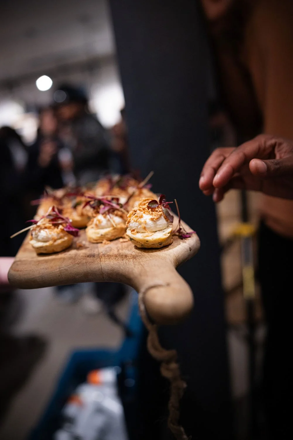 A person holding a wooden serving board with bite-sized appetizers topped with microgreens in a crowded indoor setting.