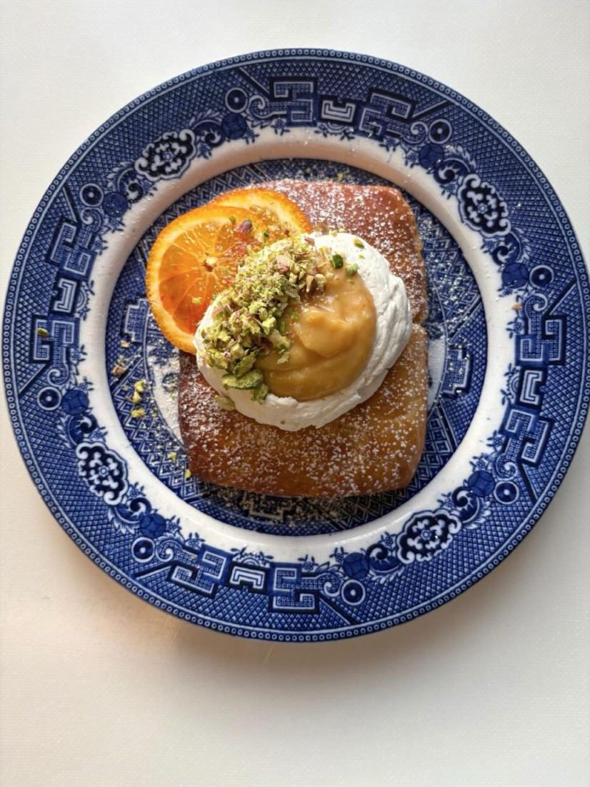 A plate with French toast topped with whipped cream, sliced orange, chopped pistachios, and a dollop of lemon curd dusted with powdered sugar.