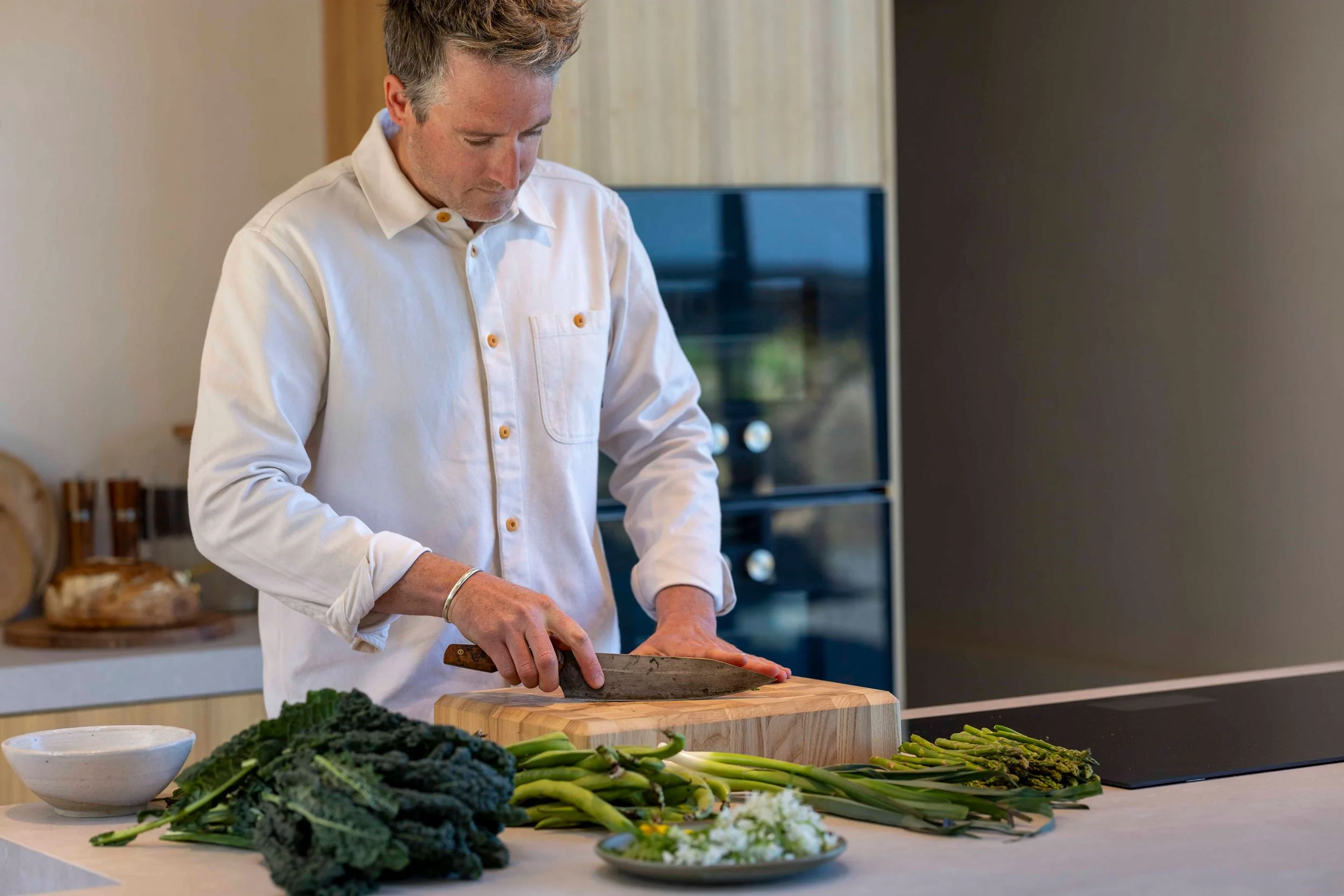 A man with gray hair, wearing a white shirt with rolled-up sleeves, is chopping vegetables on a wooden cutting board in a modern kitchen. There are various vegetables, including kale and asparagus, spread out on the kitchen counter in front of him.