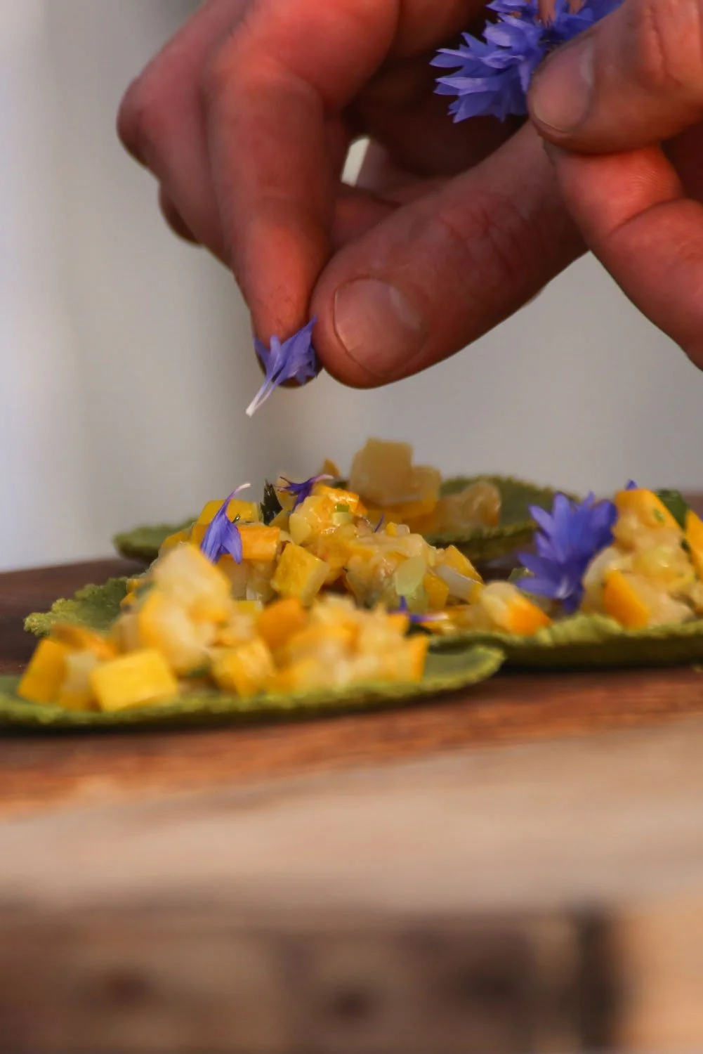 Person garnishing a dish with purple edible flowers, with a focus on their hand and the vibrant dish underneath