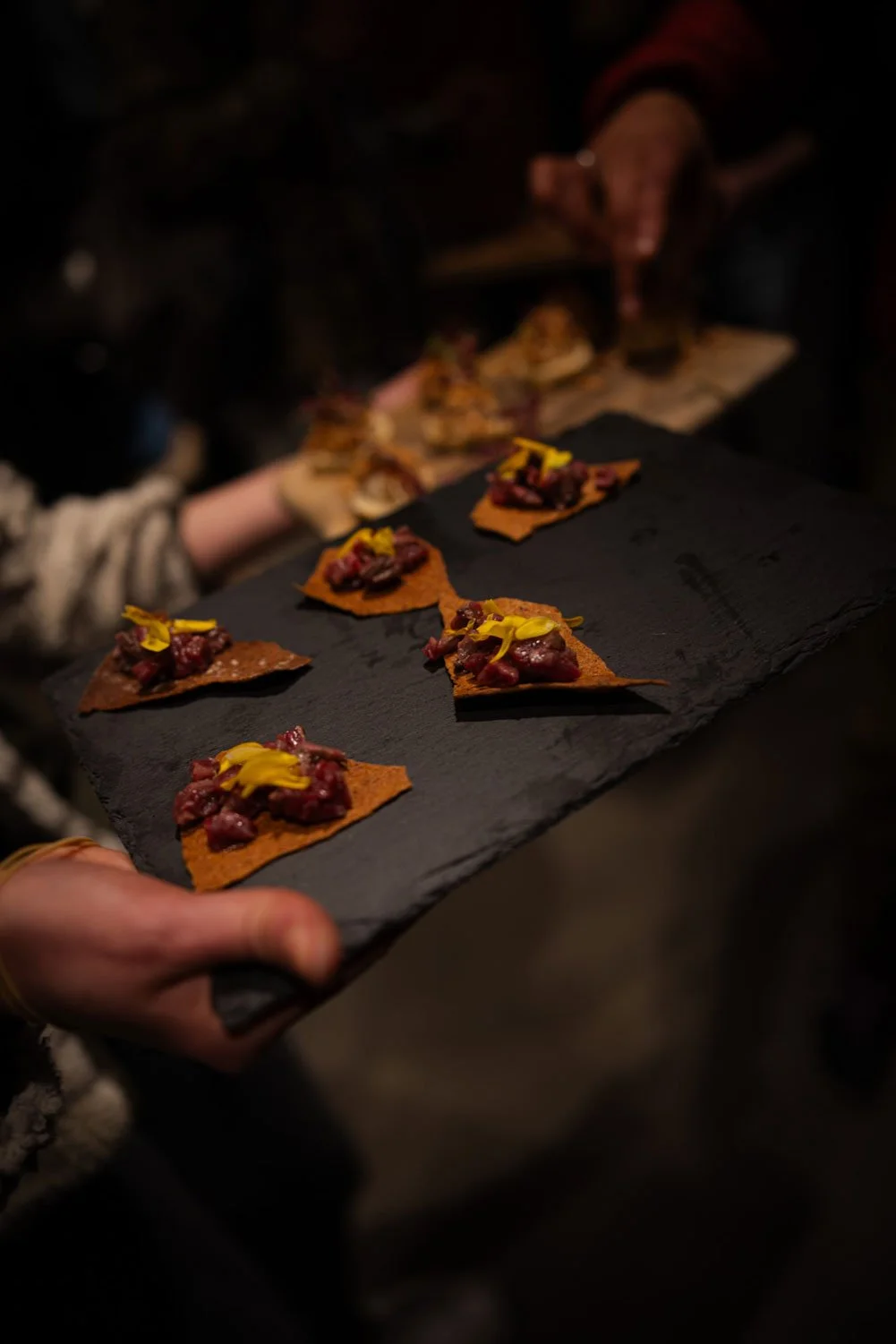 Close-up of a black slate serving platter with multiple small canapé appetizers topped with diced meat and yellow garnish, held by a person at a dimly lit event.