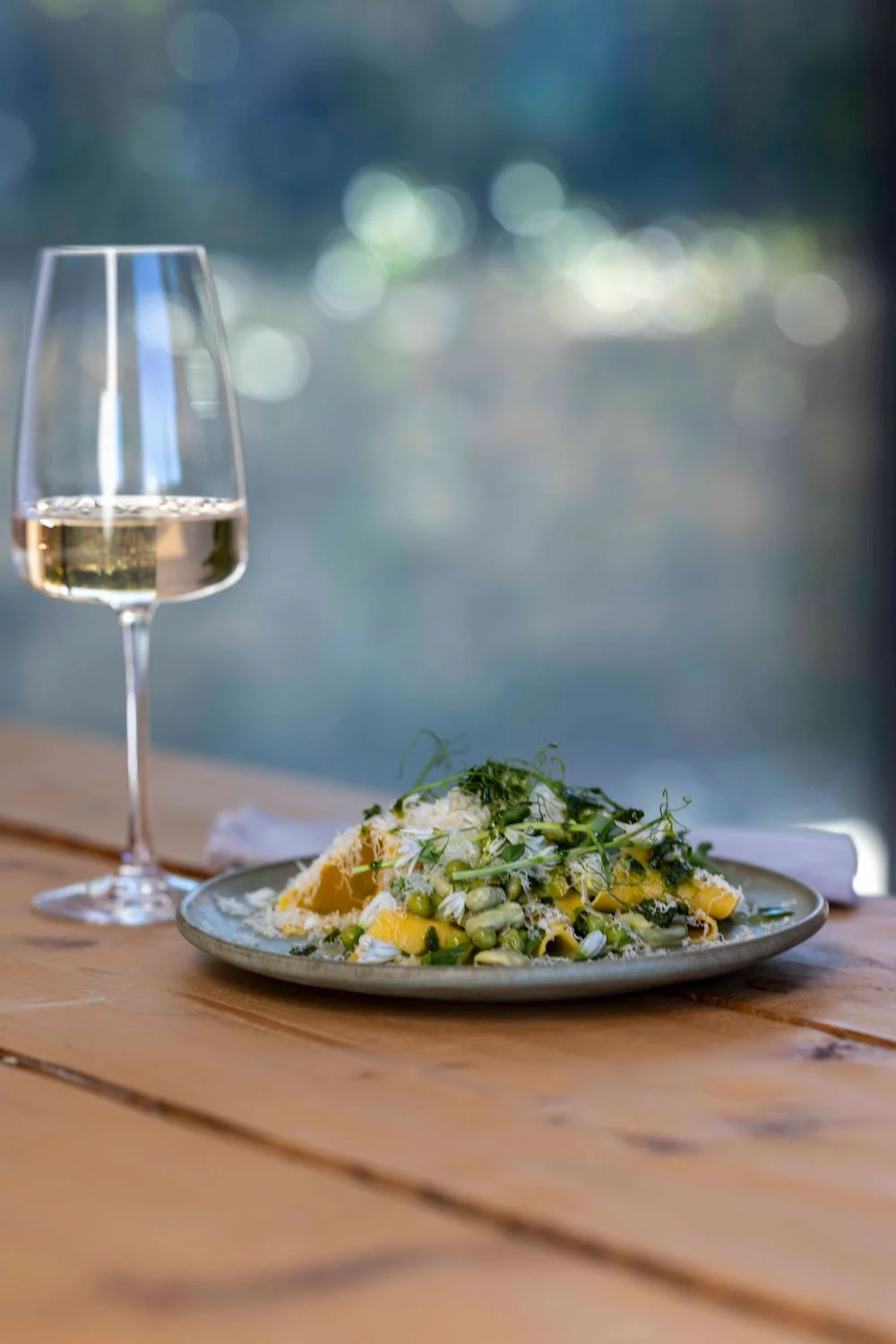 A glass of white wine and a plate of pasta with greens on a wooden table with a blurred outdoor background.