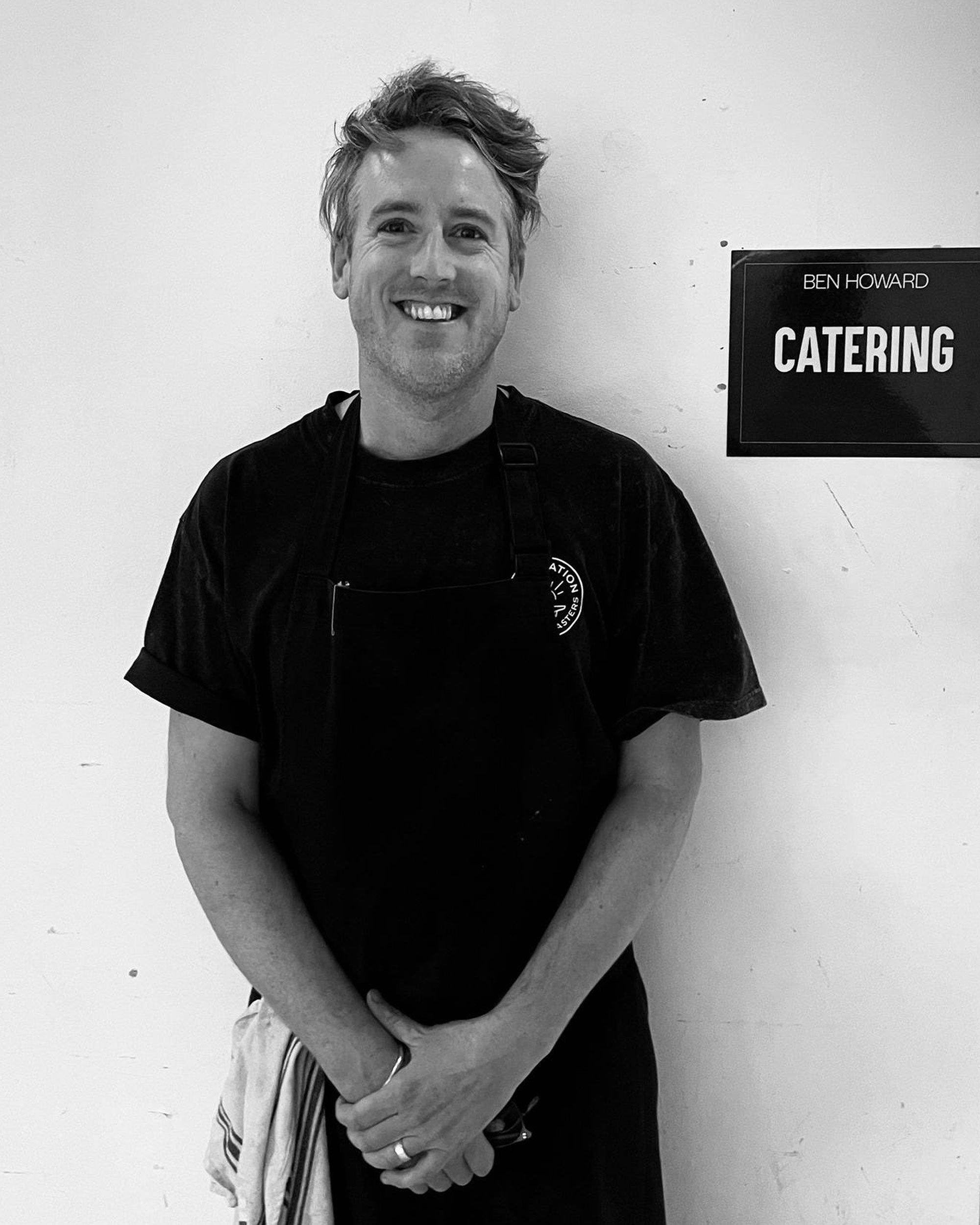 A smiling man with wavy hair, wearing a black shirt, stands against a plain wall with a sign that reads 'Ben Howard Catering'.