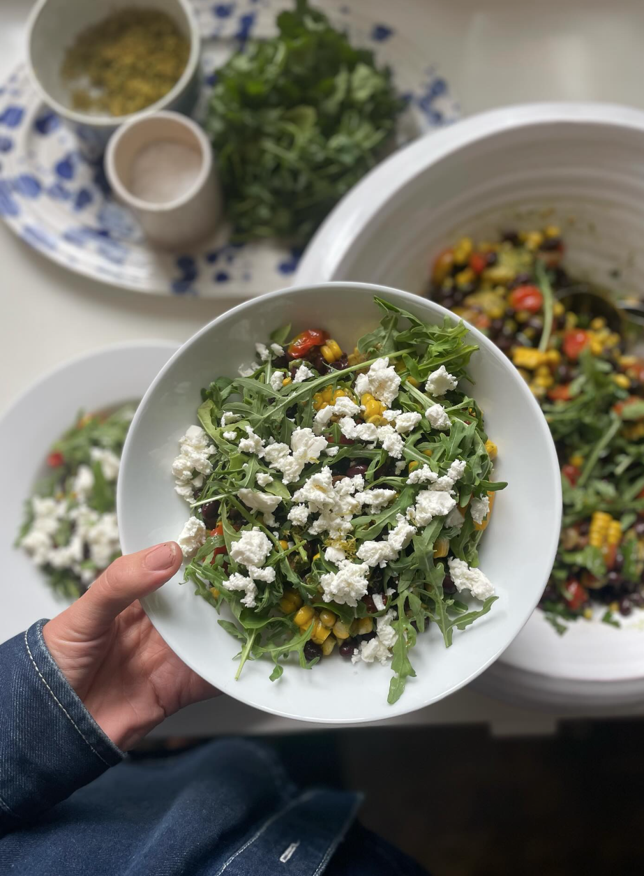 A person holding a white bowl of fresh salad with arugula, corn, cherry tomatoes, and crumbled cheese, with additional salad bowls and ingredients on a table in the background.