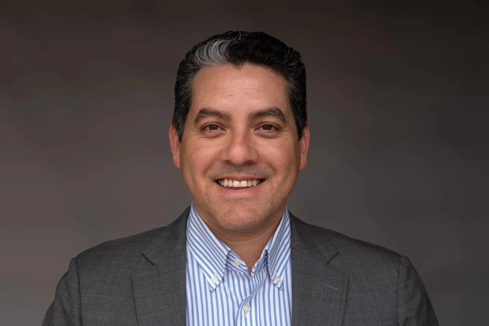 Portrait of a man with dark, combed-back hair, smiling, wearing a dark suit and a light blue striped dress shirt, against a neutral background.