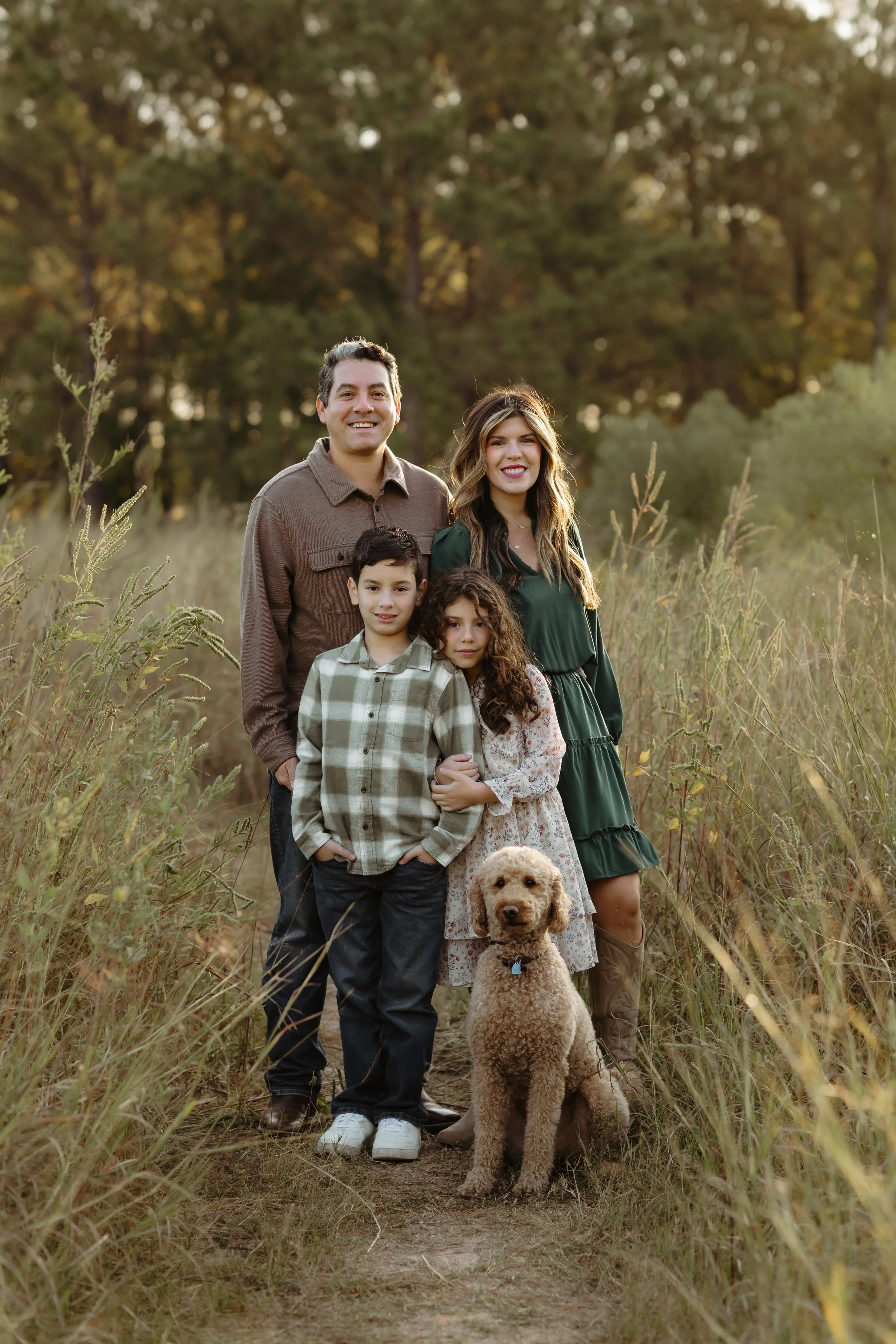 A family of five with two children and a dog standing outdoors on a dirt path in a grassy field with trees in the background, during sunset.