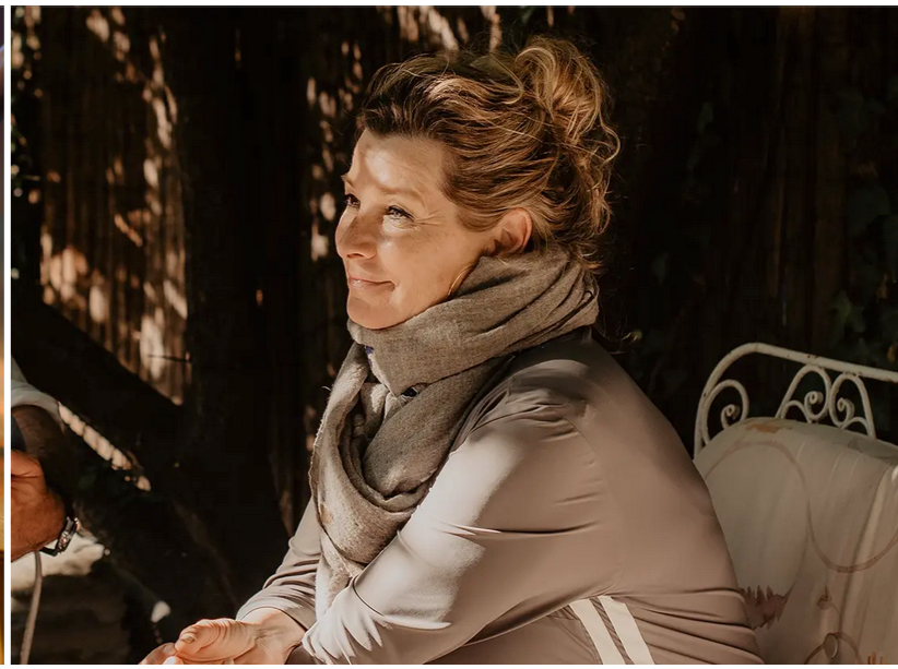Eine lächelnde Frau mit braunen, gelockten Haaren, die einen grauen Schal trägt. Sie sitzt im Freien auf einer Bank, im Hintergrund sind Schatten und eine dunkle Hecke zu sehen.