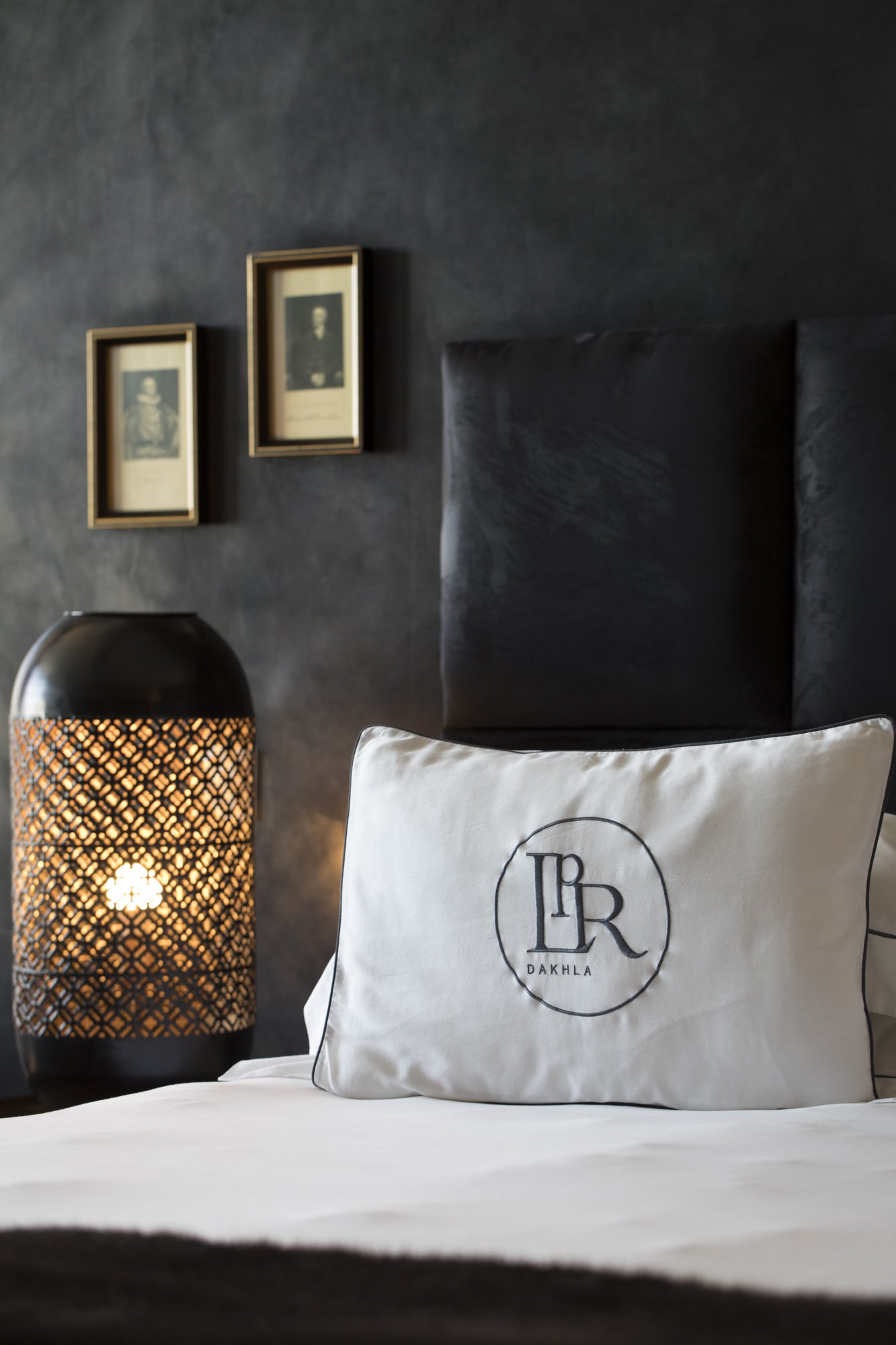 Un lit avec un coussin blanc portant le logo 'LR DAKHLA', à côté d'une lampe noire avec un motif en treillis, dans une chambre avec un mur gris foncé et deux portraits encadrés au mur.
