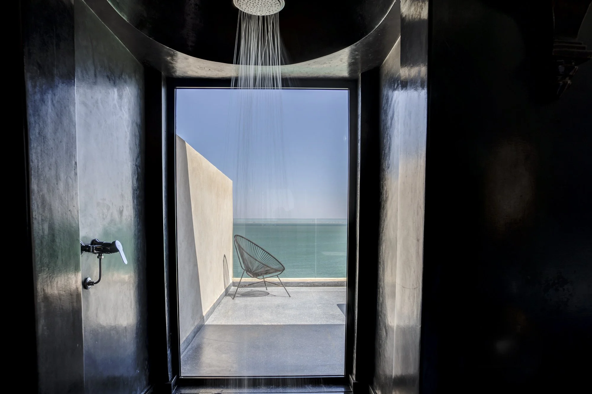 Vue depuis une salle de bain sombre vers un balcon avec une chaise en métal, vue sur la mer au loin, avec une douche en haut.