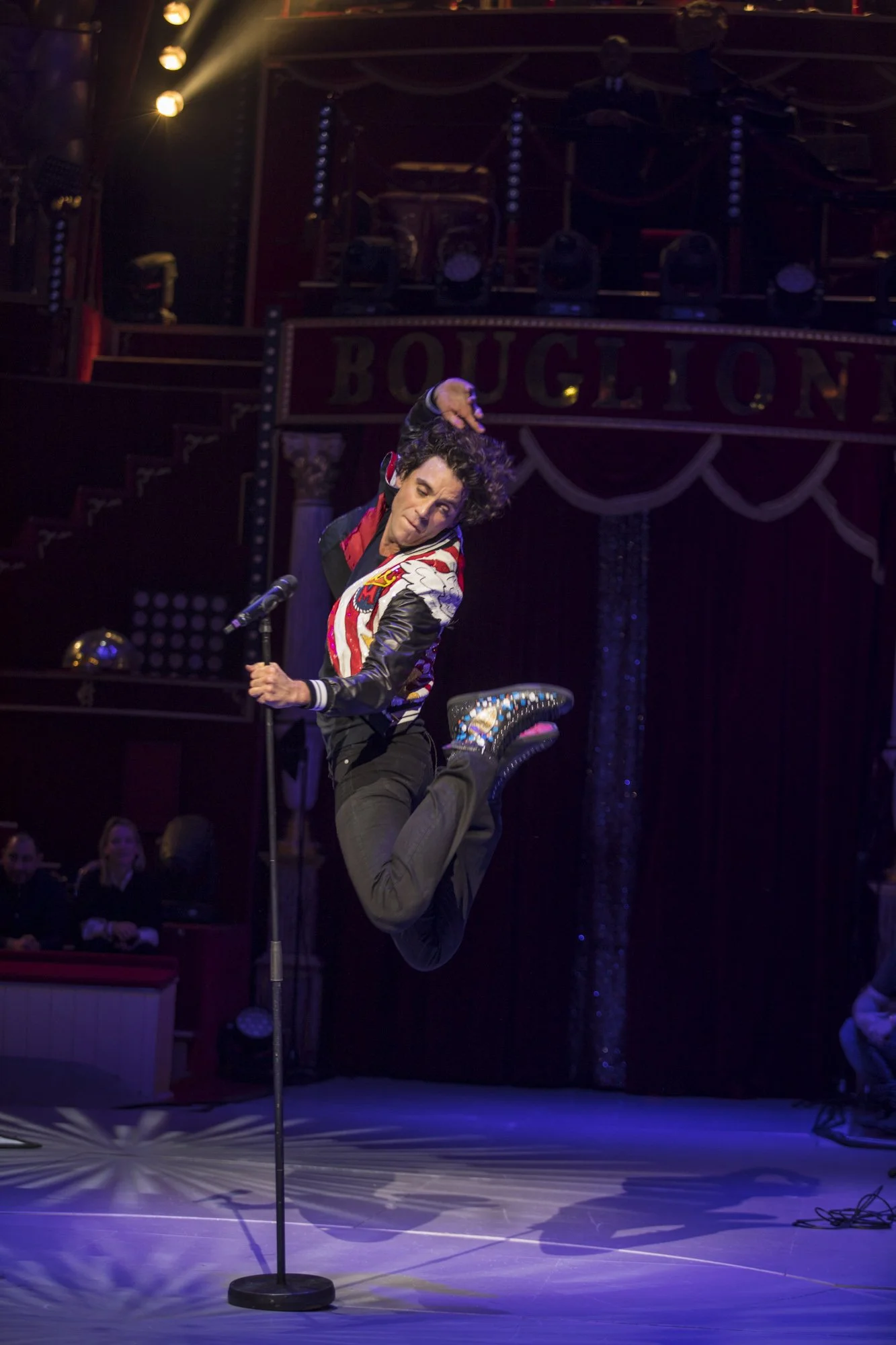 Un homme en costume coloré saute avec un micro sur scène dans un cirque, avec un public en arrière-plan.