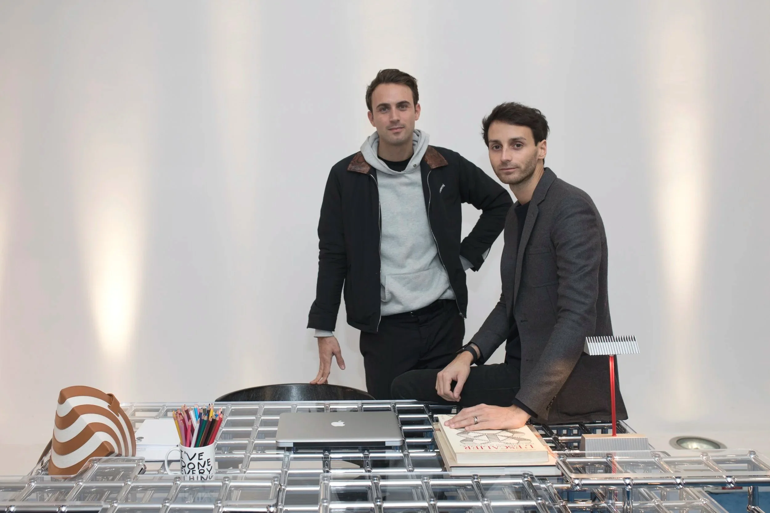 Deux hommes dans un bureau moderne, un assis sur une table en plexiglas et l'autre debout à côté. La table contient une étiquette pour stylos, un ordinateur portable, un magazine, une maquette, et un pot avec des stylos. Fond blanc.