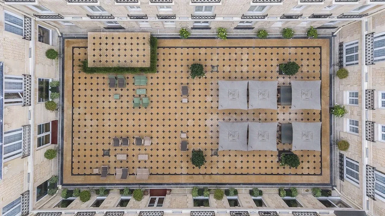Vue en hauteur d'une cour intérieure d'un immeuble avec des parasols, des chaises, des arbres et un sol en motif géométrique.