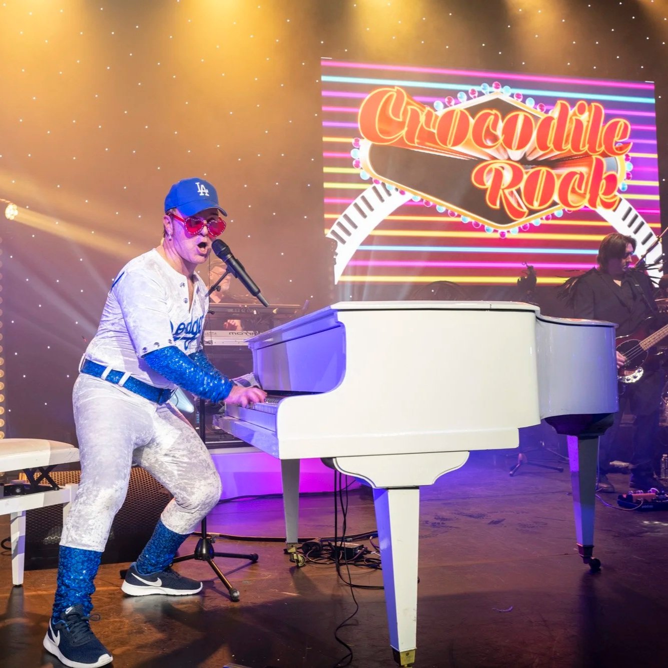 Une performance musicale sur scène avec un homme jouant du piano blanc, portant des vêtements flashy et des accessoires colorés, avec une guitare en arrière-plan. Un écran en arrière-plan affiche le texte « Crocodile Rock » en lettres lumineuses.