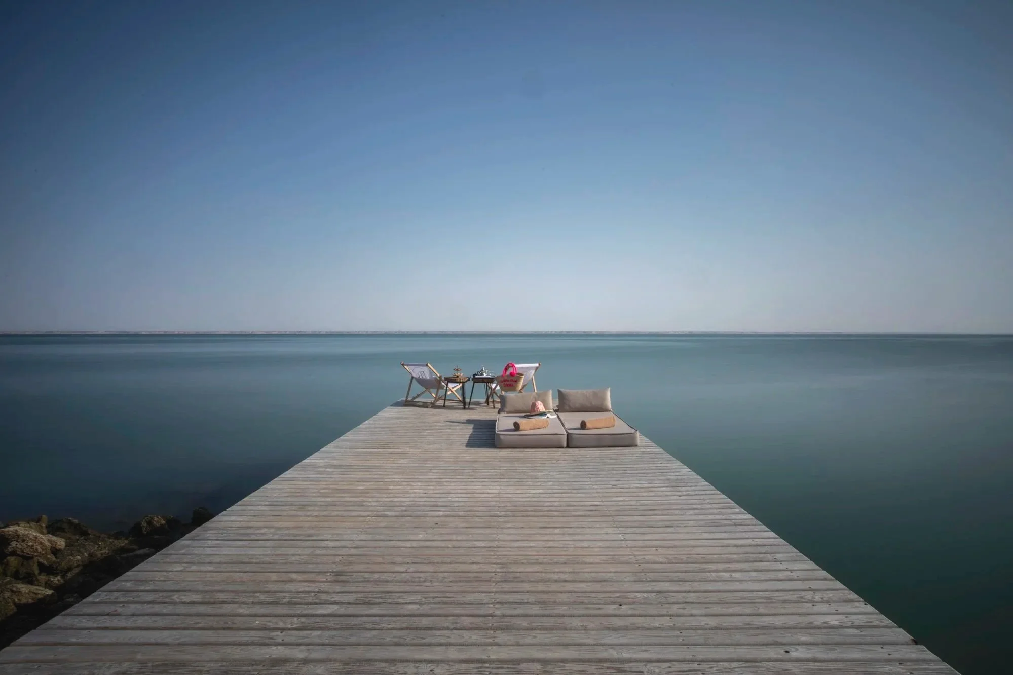 Jetée en bois flotté s'étendant sur l'eau calme avec un ciel clair et dégagé, mobilier de détente comprenant deux chaises longues, des coussins, une petite table avec des objets et des sacs à Beach à l'arrière.