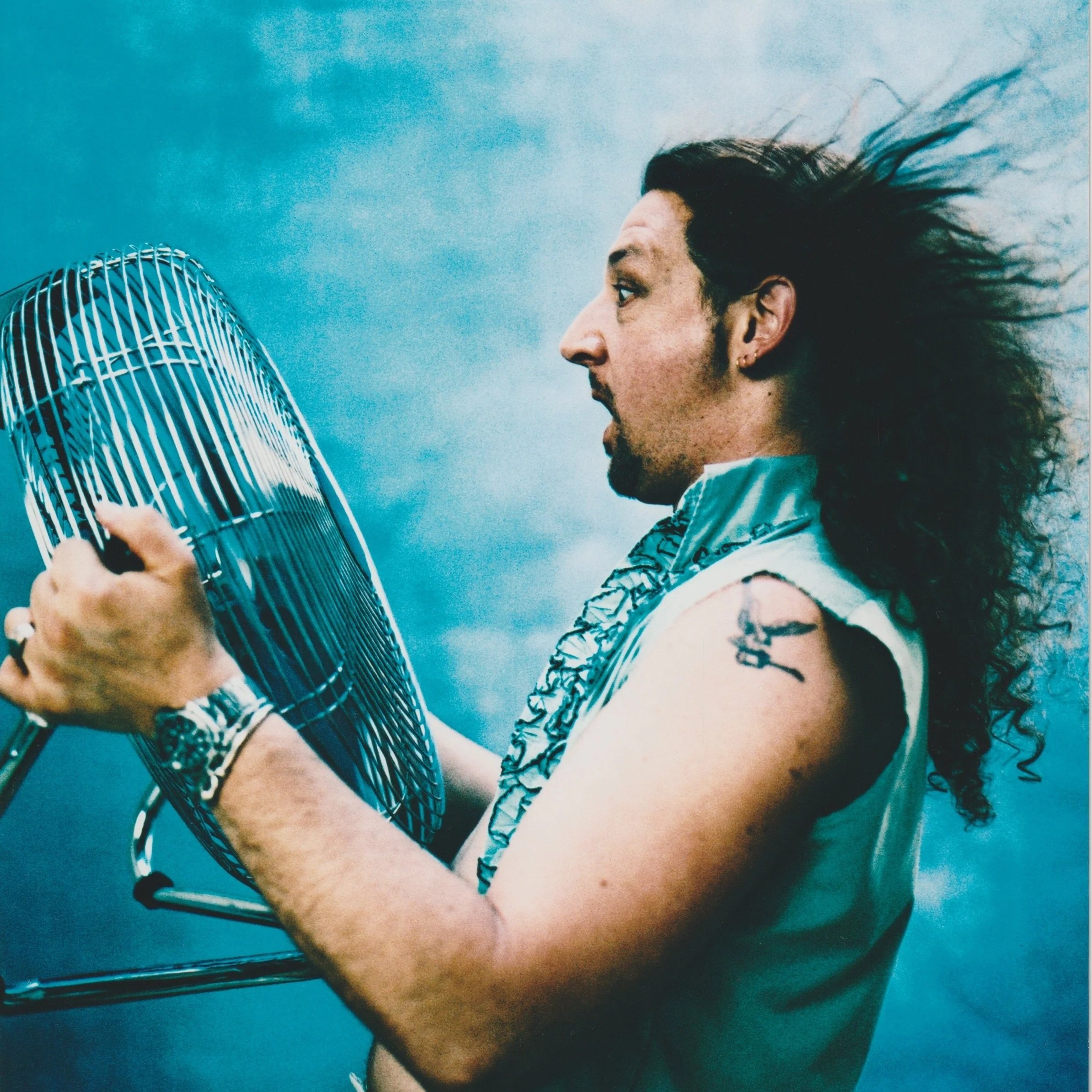 Un homme à cheveux longs et bouclés, portant un vêtement sans manches et une montre, tient un ventilateur électrique. Il a une tatouage sur le bras et porte un bracelet.