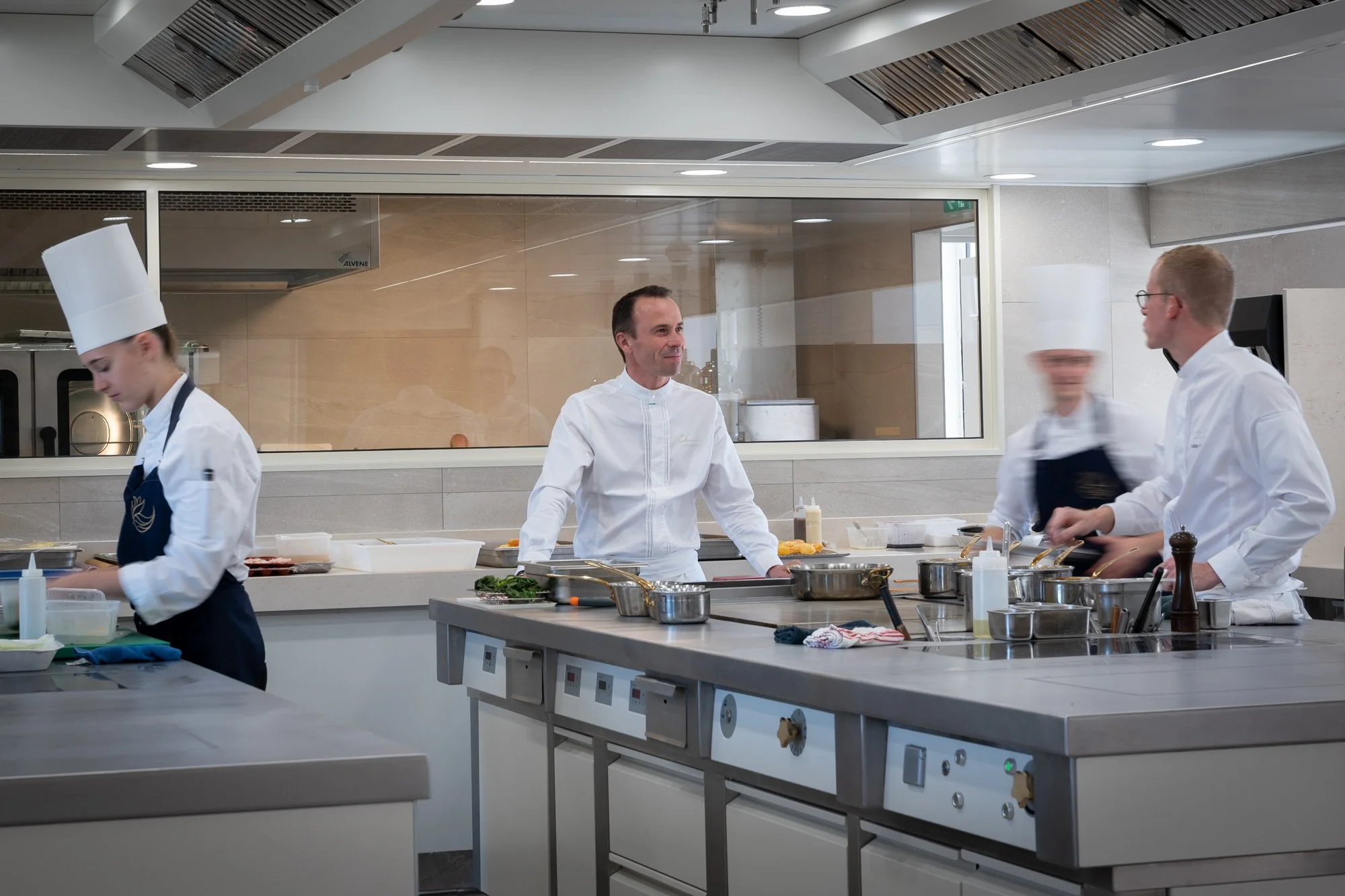 Cinq chefs en tenue de cuisine dans une cuisine professionnelle moderne, préparant et discutant autour de la zone de cuisson.