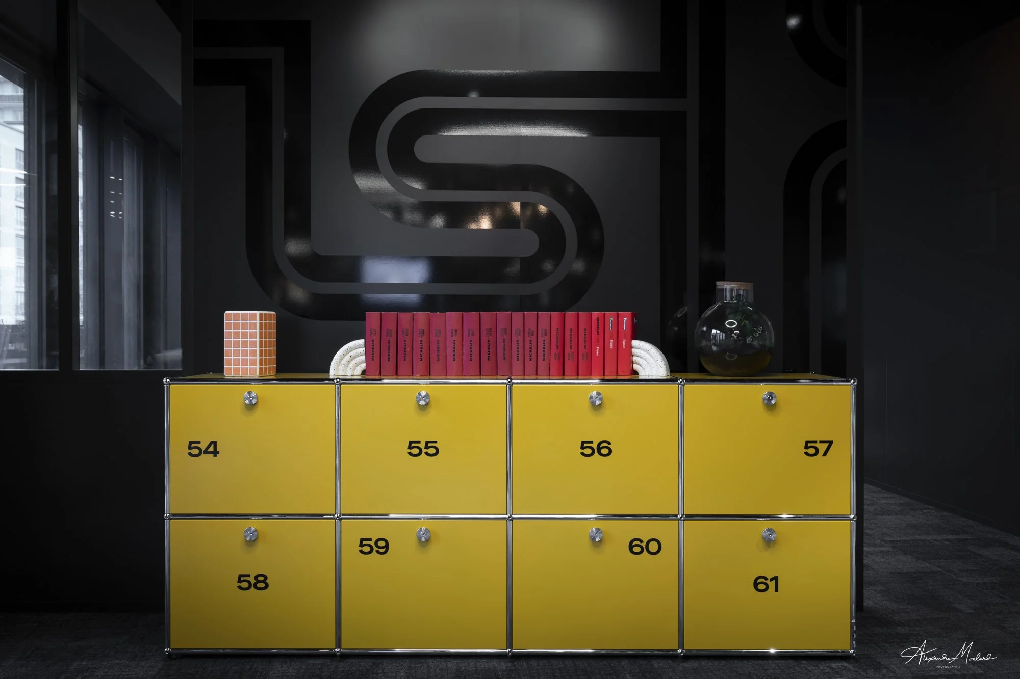 Une armoire à deux niveaux de casiers jaunes numérotés de 54 à 61, décorée avec un cube en céramique orange, un livre rouge et un verre avec une plante verte à l'intérieur, dans une pièce moderne avec un mur noir en arrière-plan.