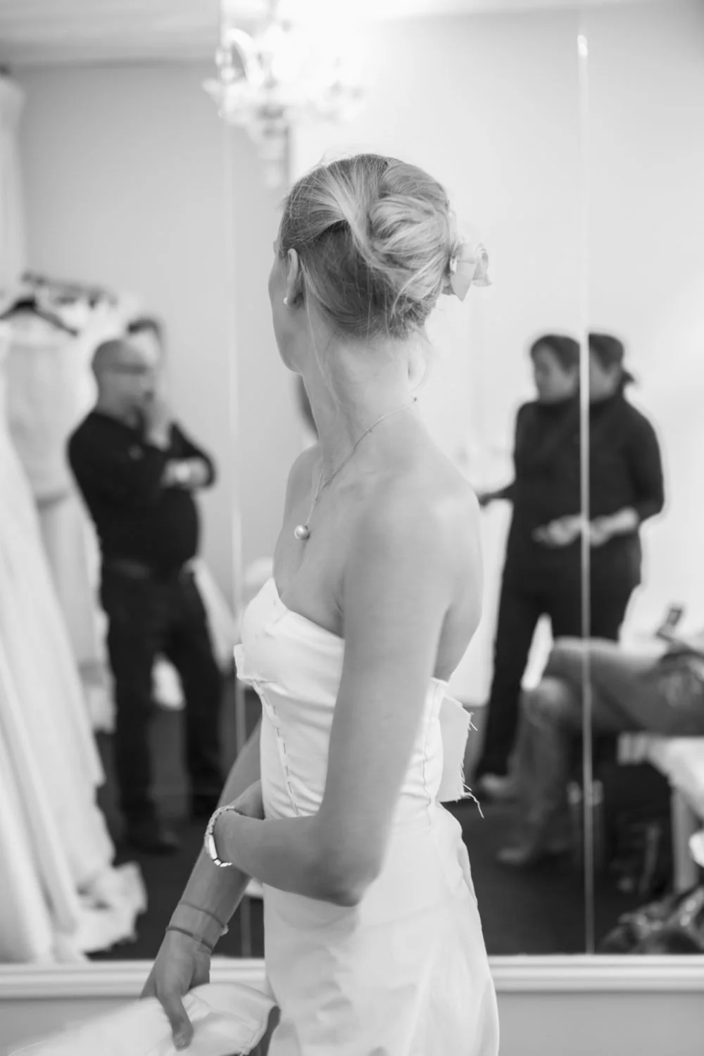 Femme en robe de mariage regardant dans un miroir, avec deux personnes floues en arrière-plan dans une pièce élégante.