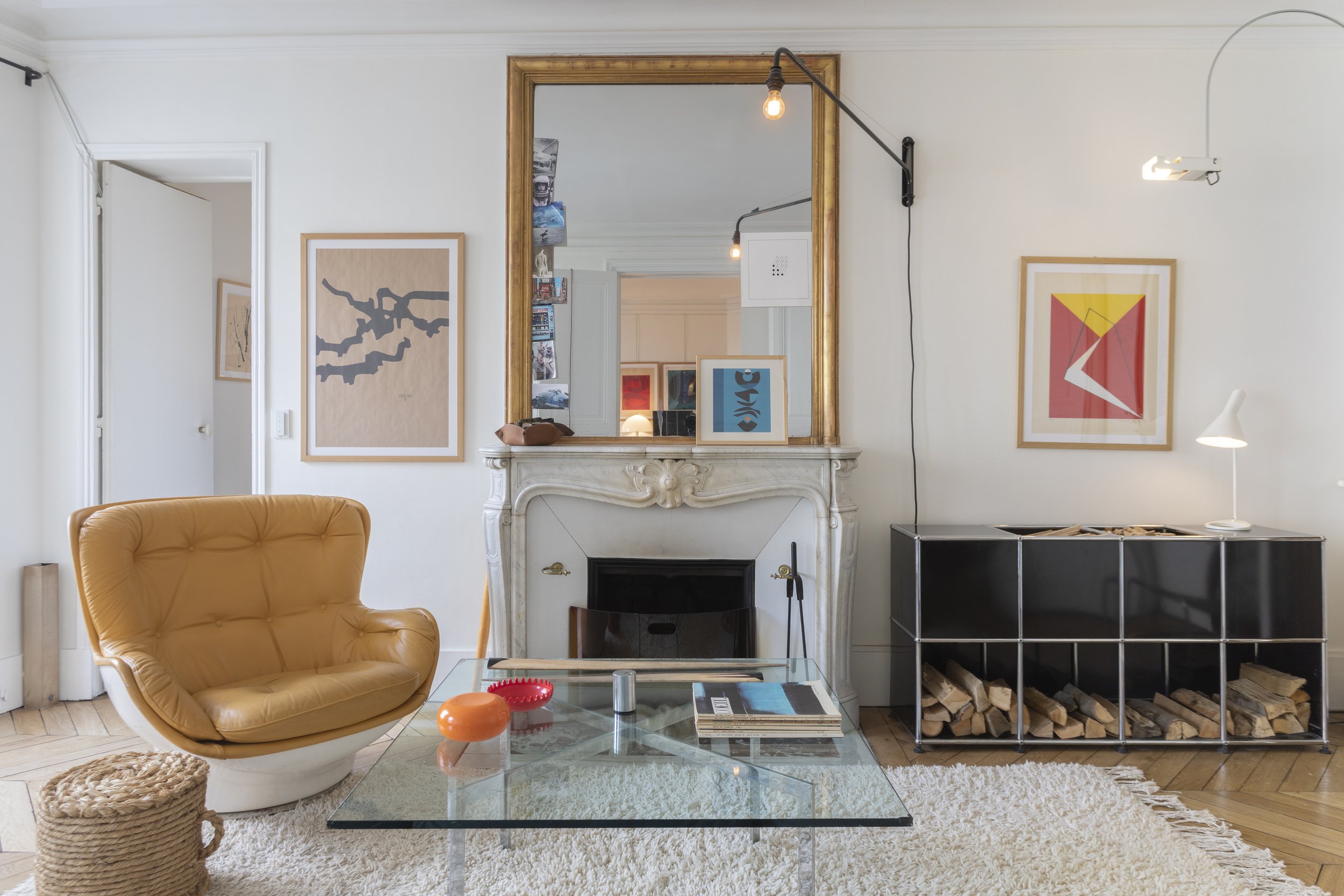 Intérieur d'un salon moderne avec un fauteuil en cuir marron, une cheminée en marbre blanc avec un grand miroir doré au-dessus, une étagère en métal contenant du bois de chauffage, des œuvres d'art sur les murs, et une table en verre avec des objets 