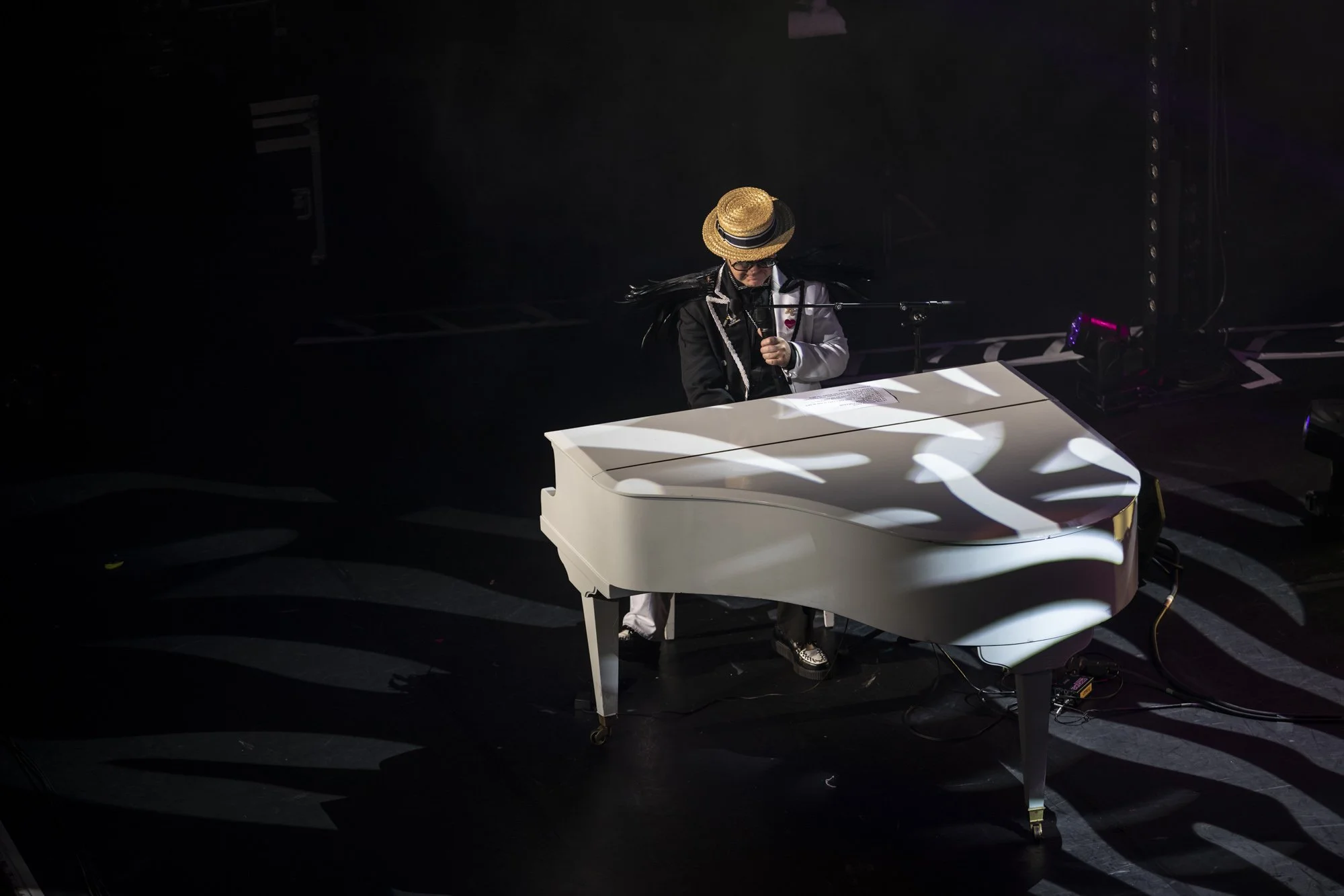 Une personne portant un chapeau de paille, des lunettes noires, et un costume noir et blanc joue du piano blanc sur scène, éclairée par un éclairage dramatique.