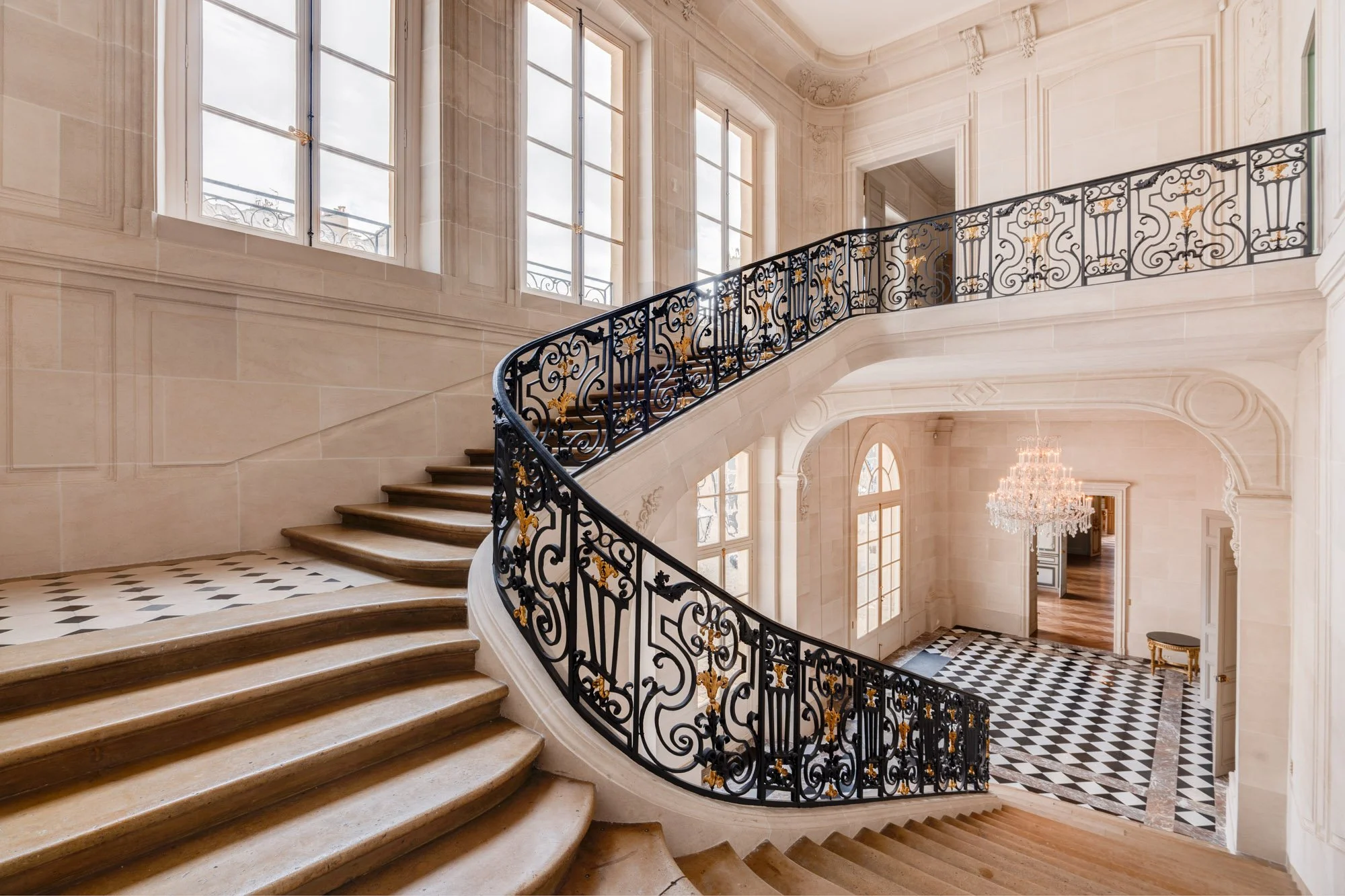 Escalier en marbre avec rampe en fer orné de détails en or, dans un intérieur élégant avec grandes fenêtres et lustre suspendu.