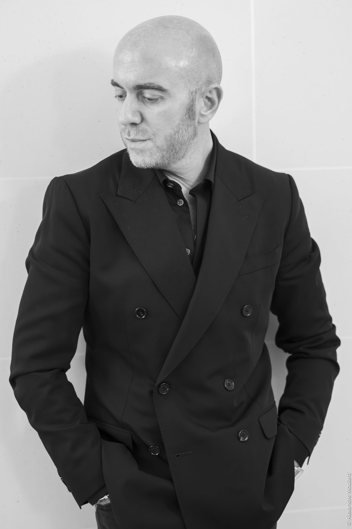 Homme en costume noir, la tête inclinée, regardant vers le haut, portant une montre, en arrière-plan blanc.