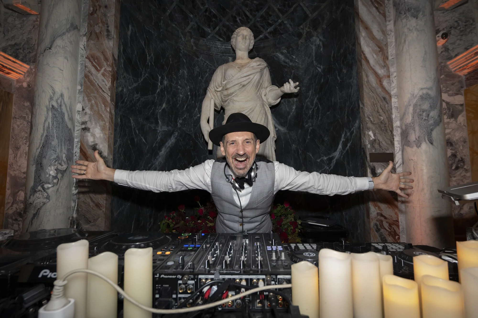 Un DJ souriant avec un chapeau noir, des écouteurs autour du cou, jouant de la musique derrière un platine DJ, avec une statue antique en marbre derrière lui dans une ambiance élégante.