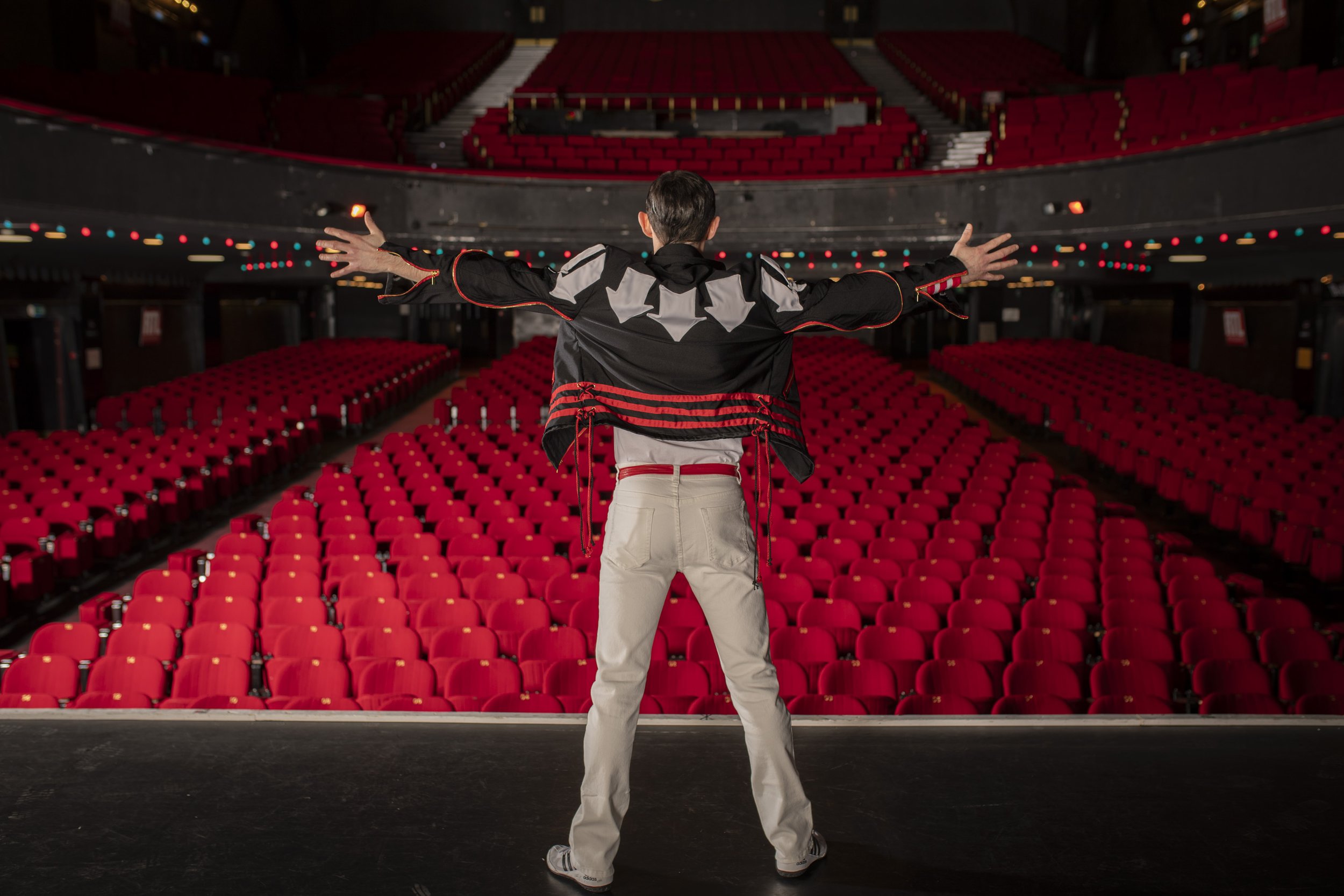 Un homme debout sur scène, vu de dos, avec des bras ouverts, dans une salle de théâtre vide avec des sièges rouges.