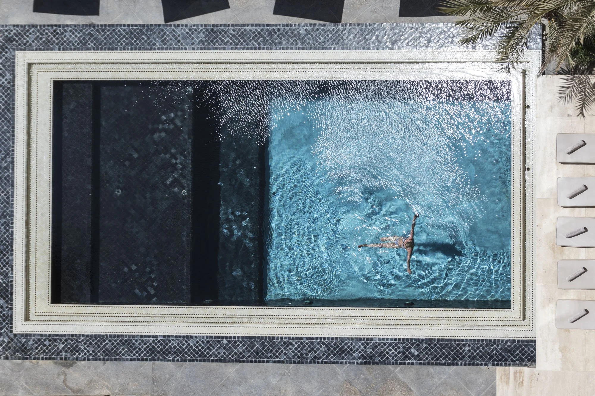 Une personne nage dans une piscine rectangulaire vue du haut, entourée de meubles et de palmiers.