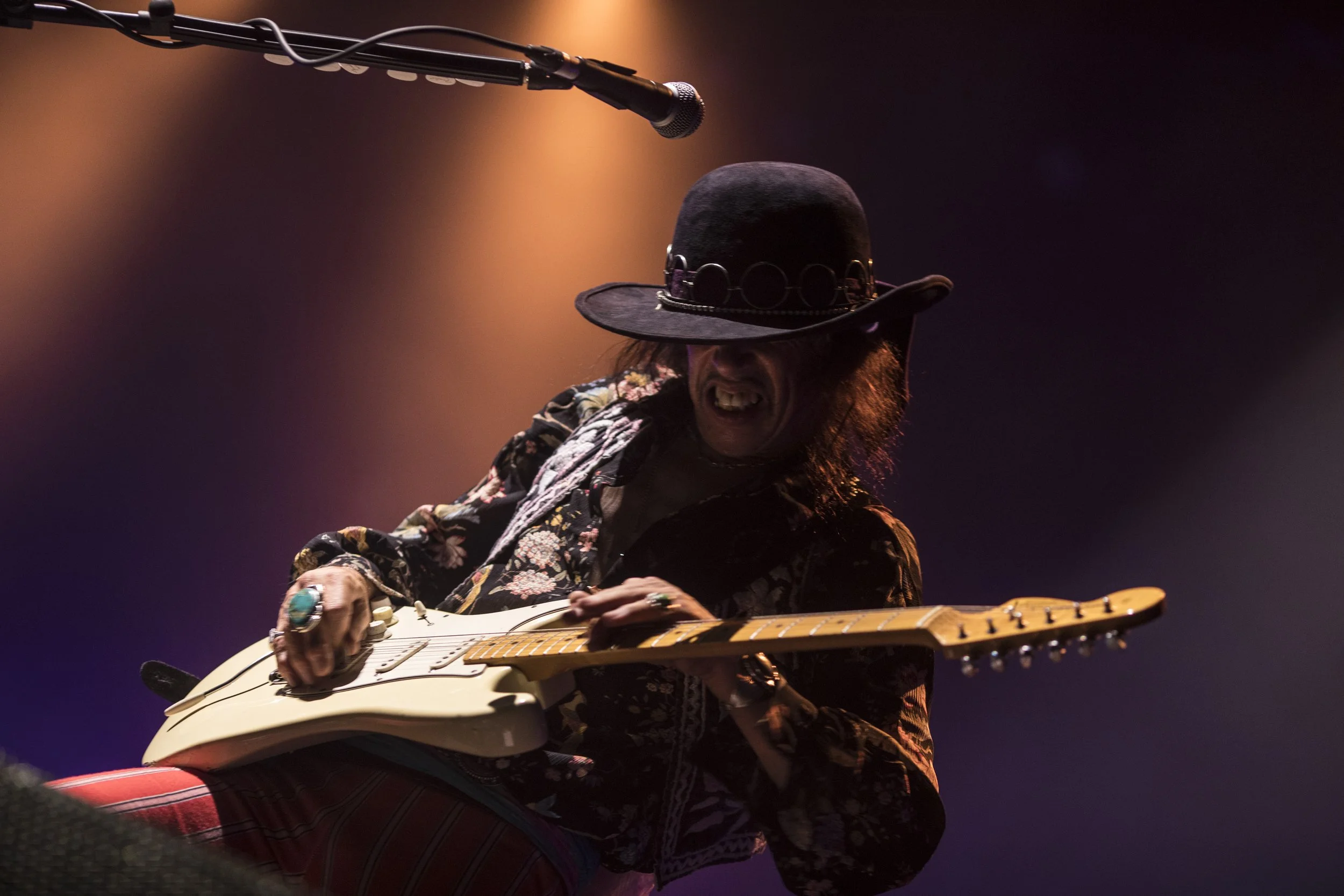 Un musicien joue de la guitare électrique sur scène, portant un chapeau noir et une chemise à motifs floraux, sous un éclairage chaud et sombre.