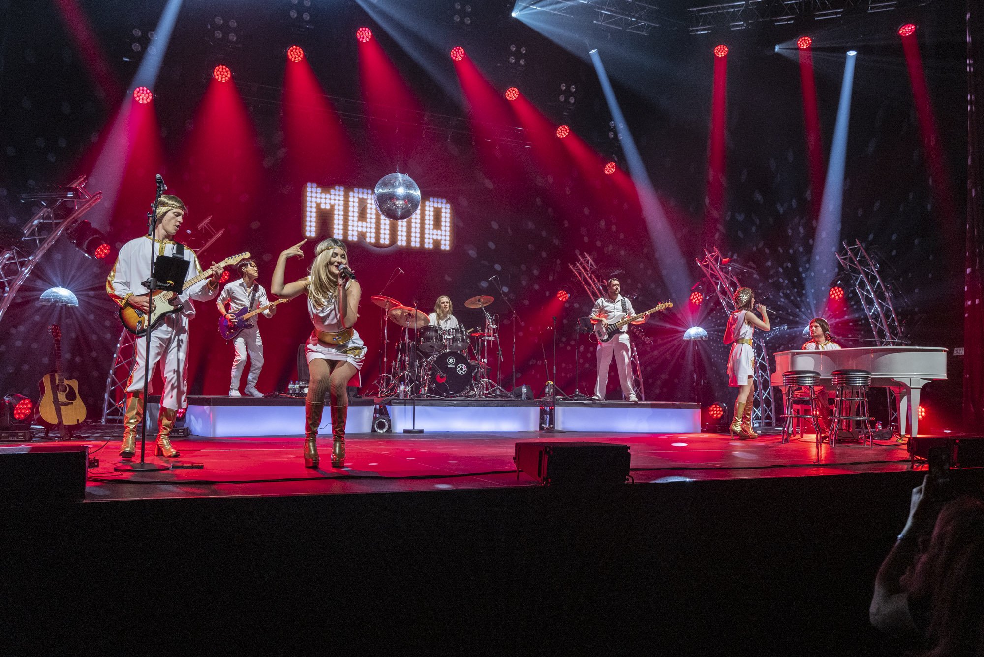 Groupe de musiciens sur scène avec éclairages rouges et blancs, une image de disque suspendue, le mot "FIASIA" affiché en lumière, et un piano blanc à droite.
