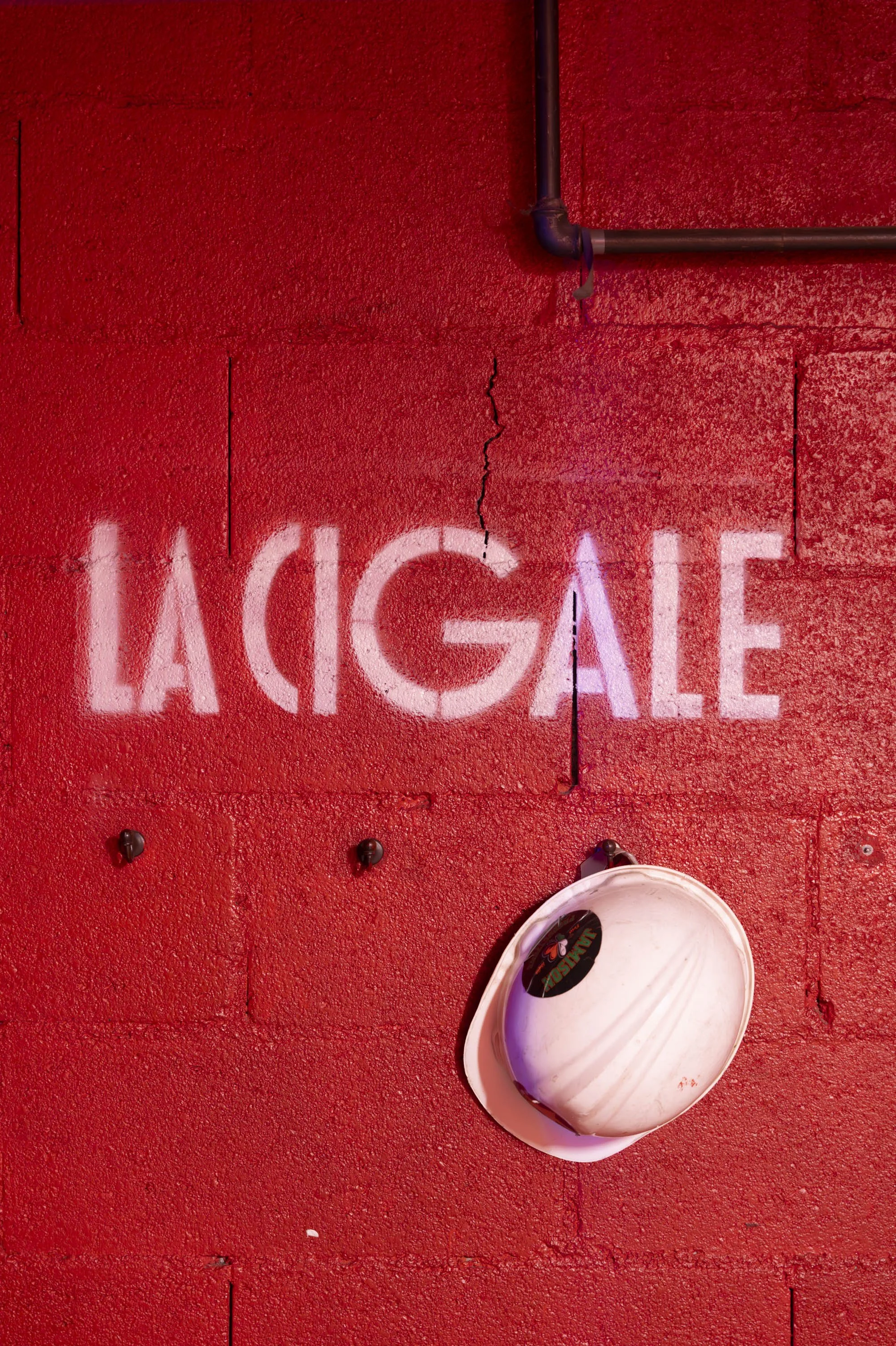 Mur rouge avec un écriteau en néon blanc disant "La Cage" et un casque de sécurité blanc suspendu.