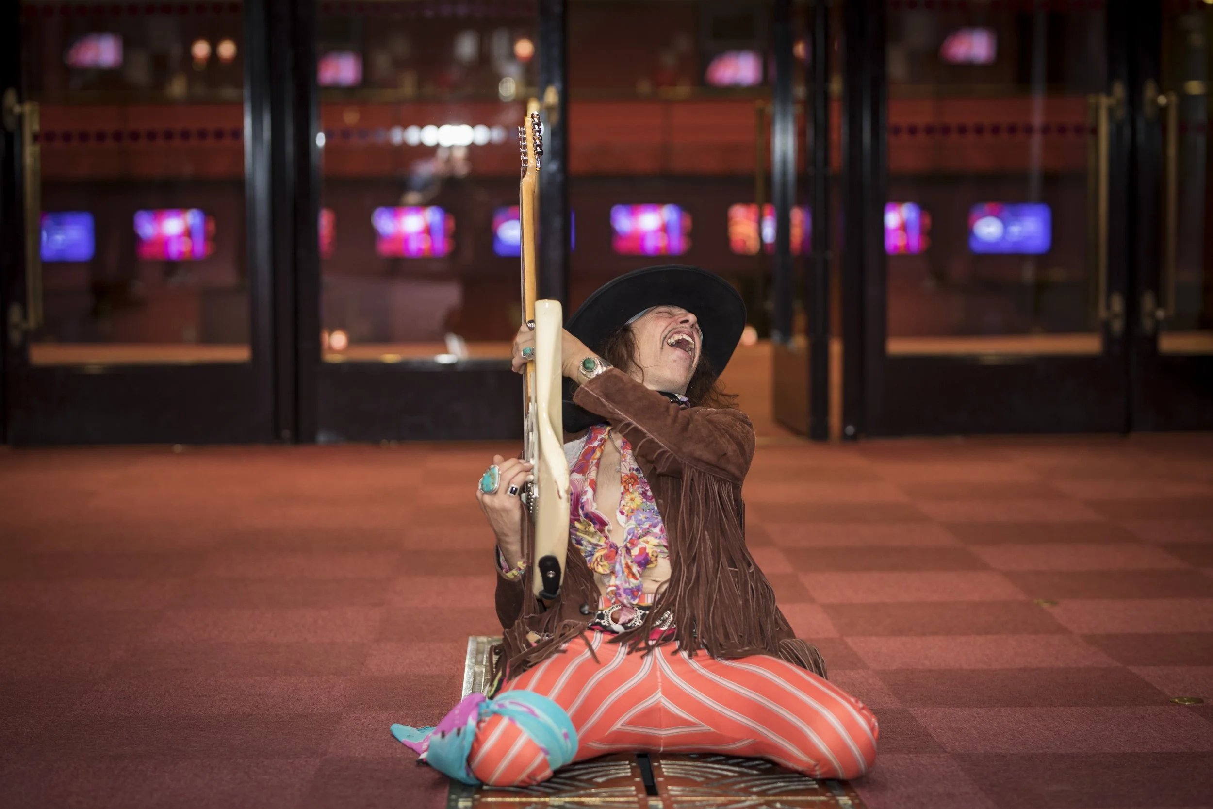 Une femme déguisée en musicien rock, assise sur le sol, jouant de la guitare électrique, cheveux longs, chapeau noir, portant un foulard coloré, veste en cuir marron avec franges, pantalon à rayures orange et blanc, bracelets et bagues turquoise, dan