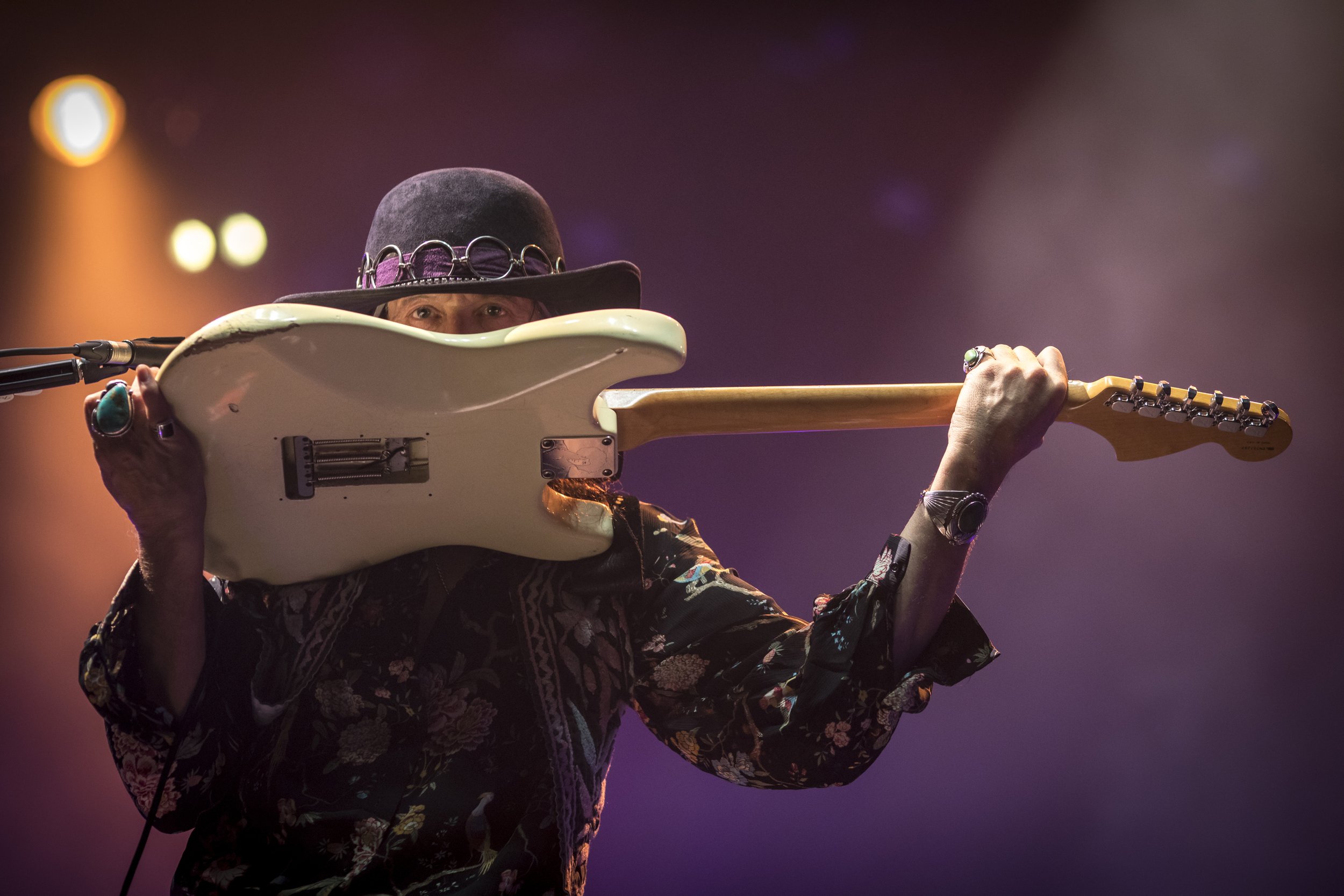 Musicien jouant de la guitare électrique sur scène, portant un chapeau noir avec des cercles en métal, décoratif, et des bijoux. Éclairage de scène coloré en arrière-plan.
