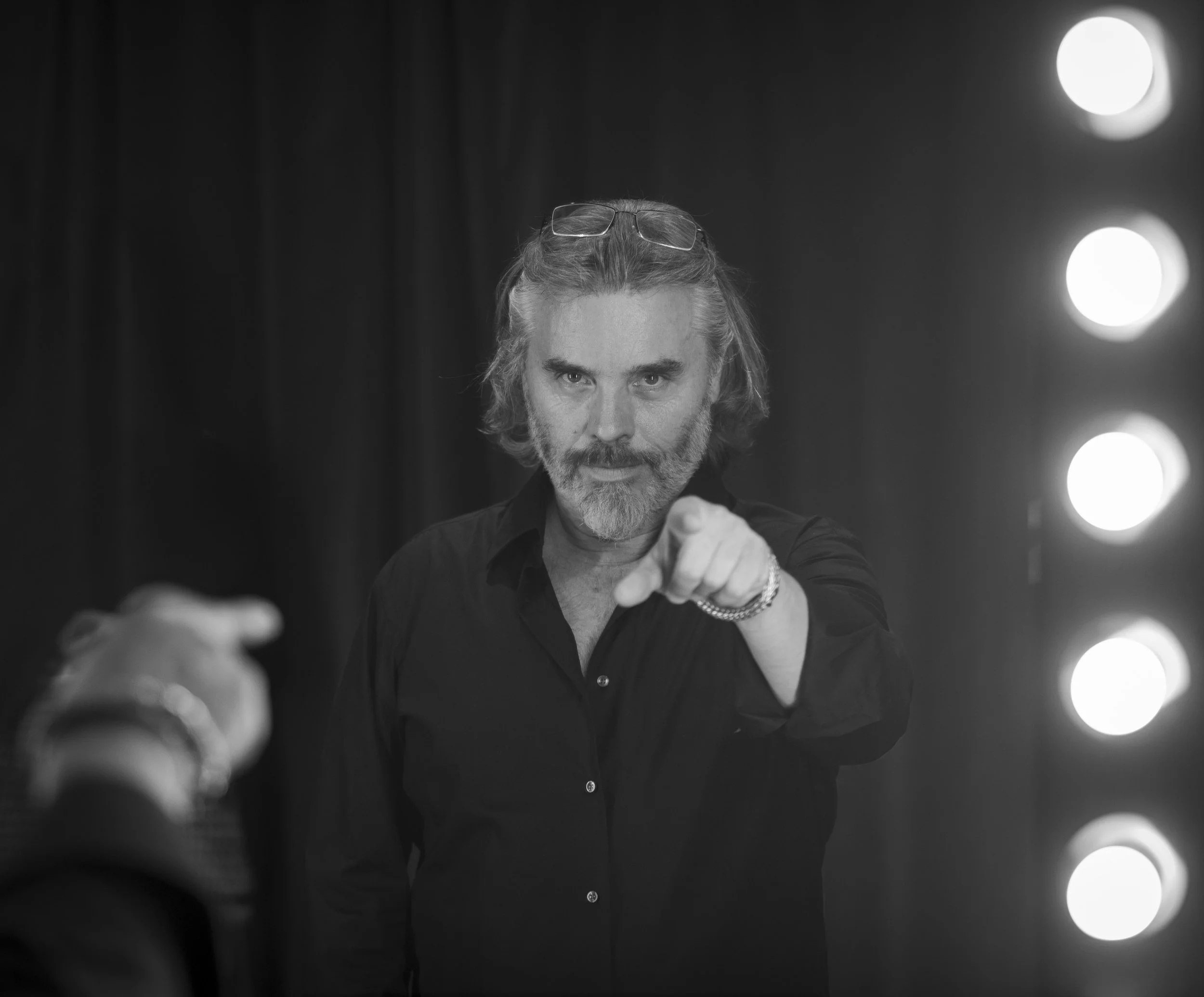 Un homme avec des cheveux longs et une barbe, portant des lunettes sur la tête, regarde dans un miroir avec un regard sérieux, pointant du doigt vers la caméra. L'éclairage est sombre avec des lumières de scène en arrière-plan.