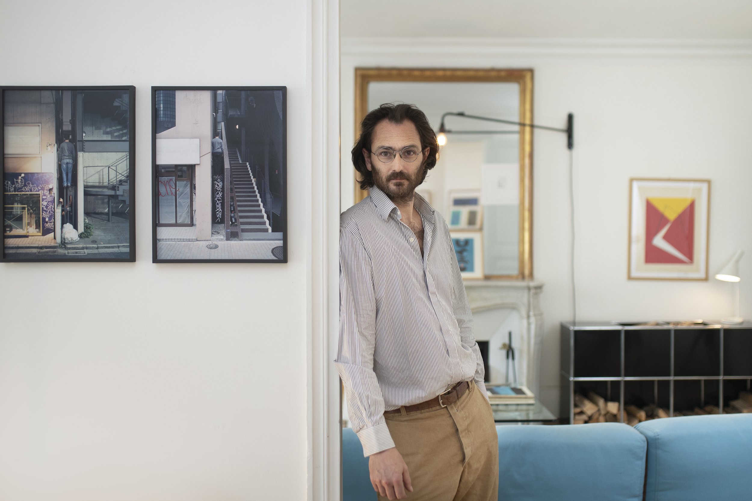 Un homme avec des lunettes, une barbe et des cheveux longs, portant une chemise à rayures et un pantalon beige, pose à côté d'une porte dans un salon moderne décoré d'œuvres d'art et de miroirs.