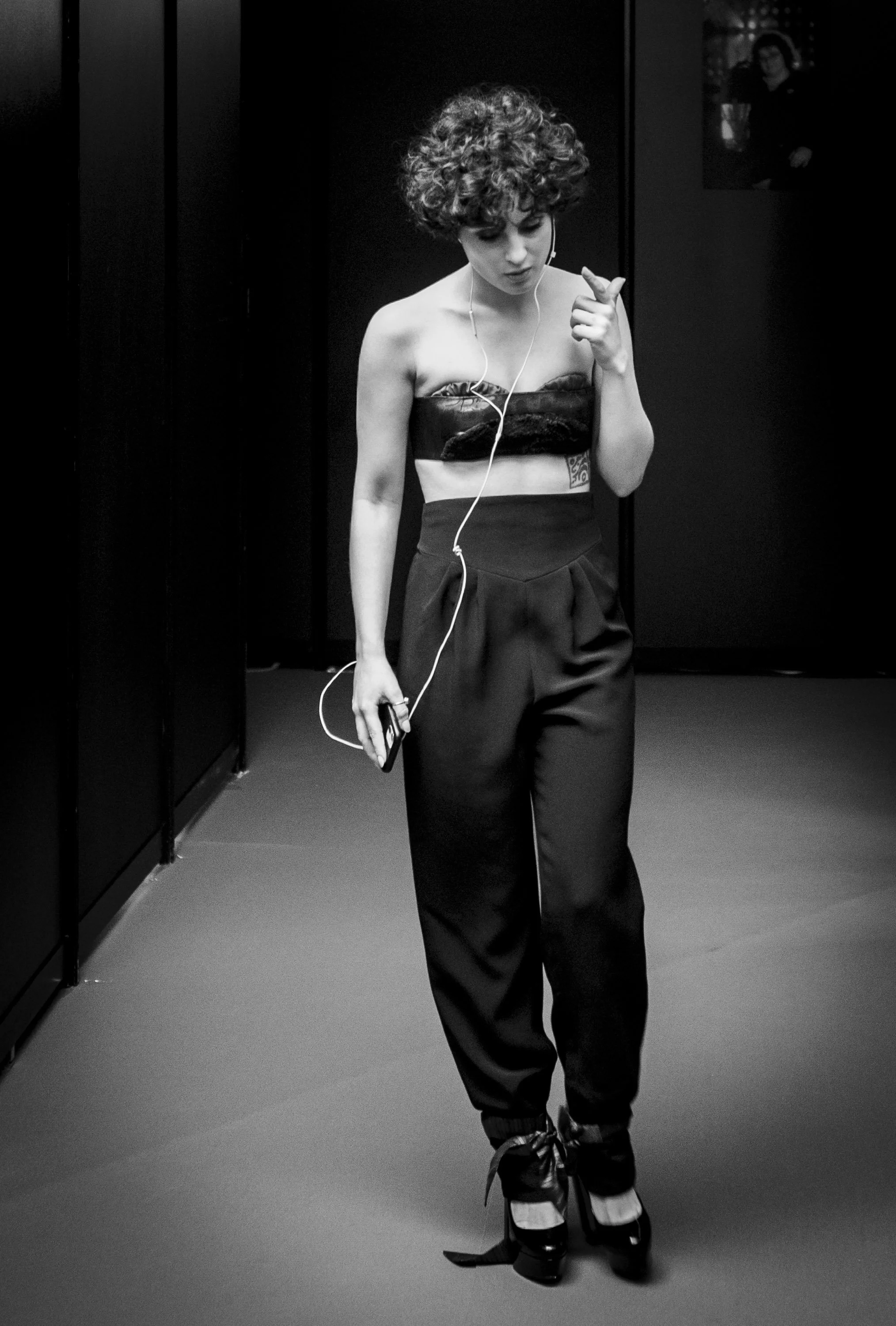 Une femme avec des cheveux bouclés courts, portant un haut court en tissu noir, un pantalon ample, et des chaussures à talons, marche dans un environnement sombre, probablement une rue ou un couloir, écoutant de la musique avec des écouteurs.