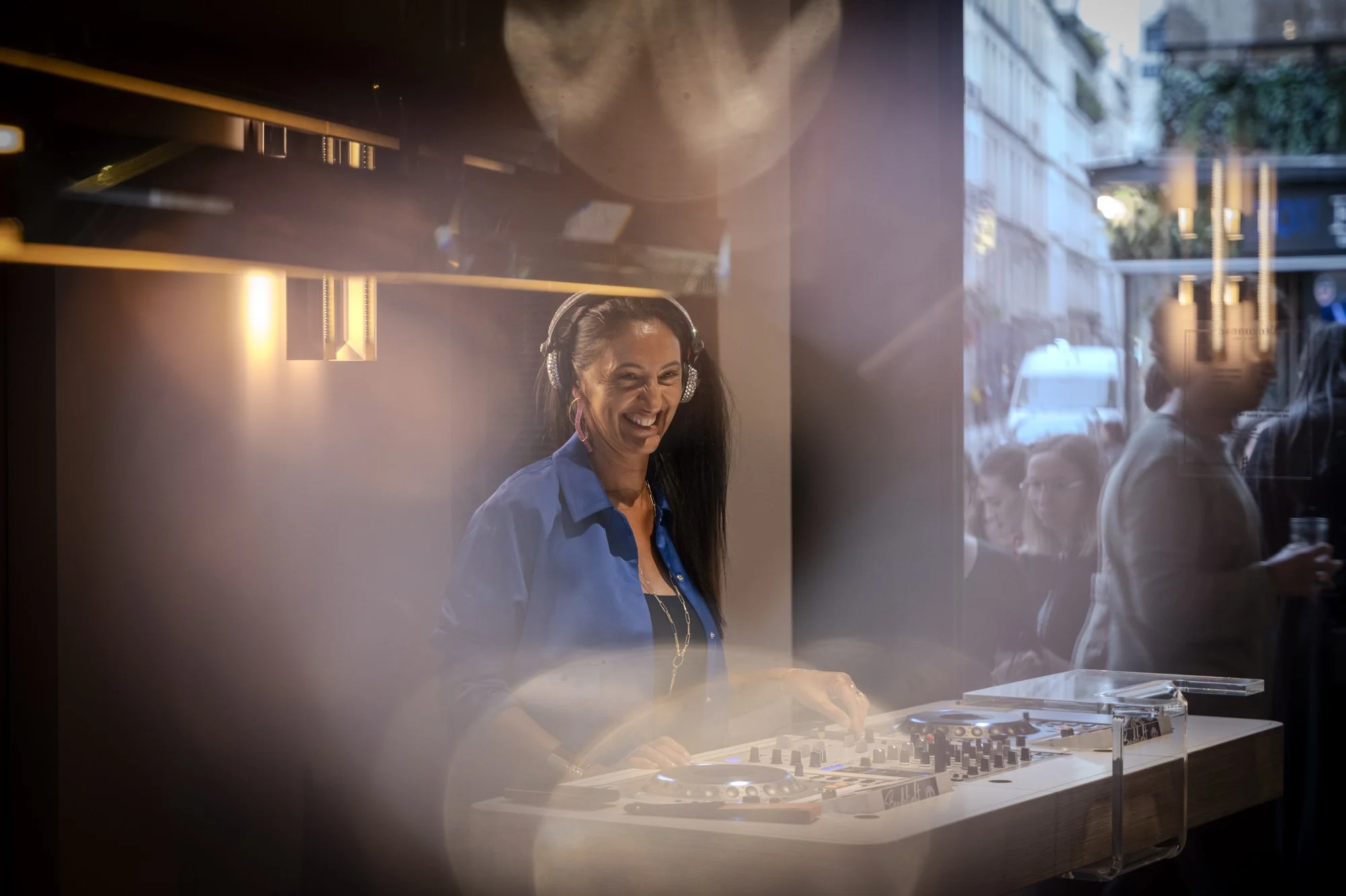Femme DJ souriante portant un casque, jouant de la musique avec un équipement DJ, vue à travers une vitre lors d'une fête en plein air avec des spectateurs et un environnement urbain en arrière-plan.