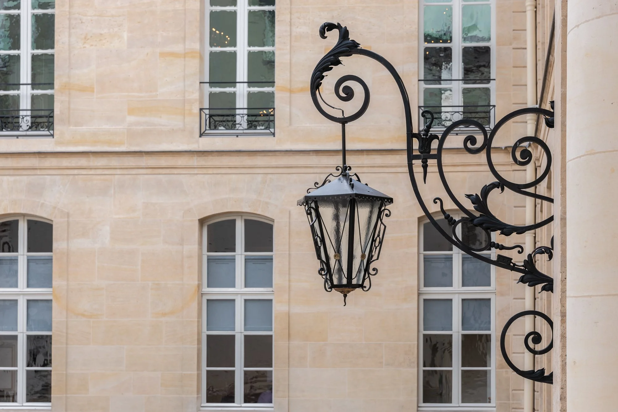 Lustre en fer forgé noir suspendu à une façade en pierre claire d'un bâtiment avec plusieurs fenêtres à huisseries blanches.