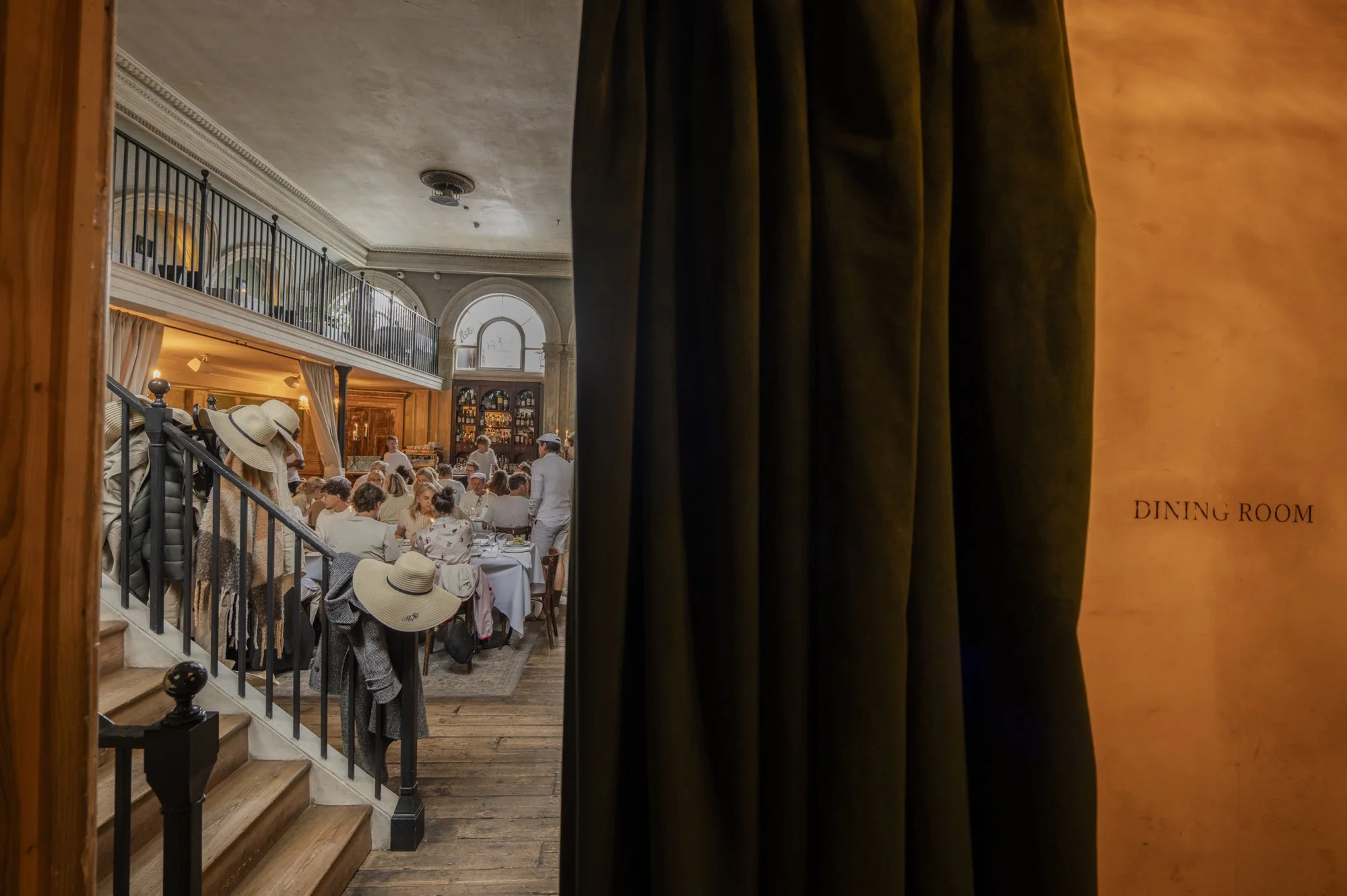 Vue à travers un rideau noir d'une salle à manger animée dans un restaurant élégant, avec des gens assis autour de tables, des serveurs en uniforme blanc et un décor classique, y compris un bar en arrière-plan.