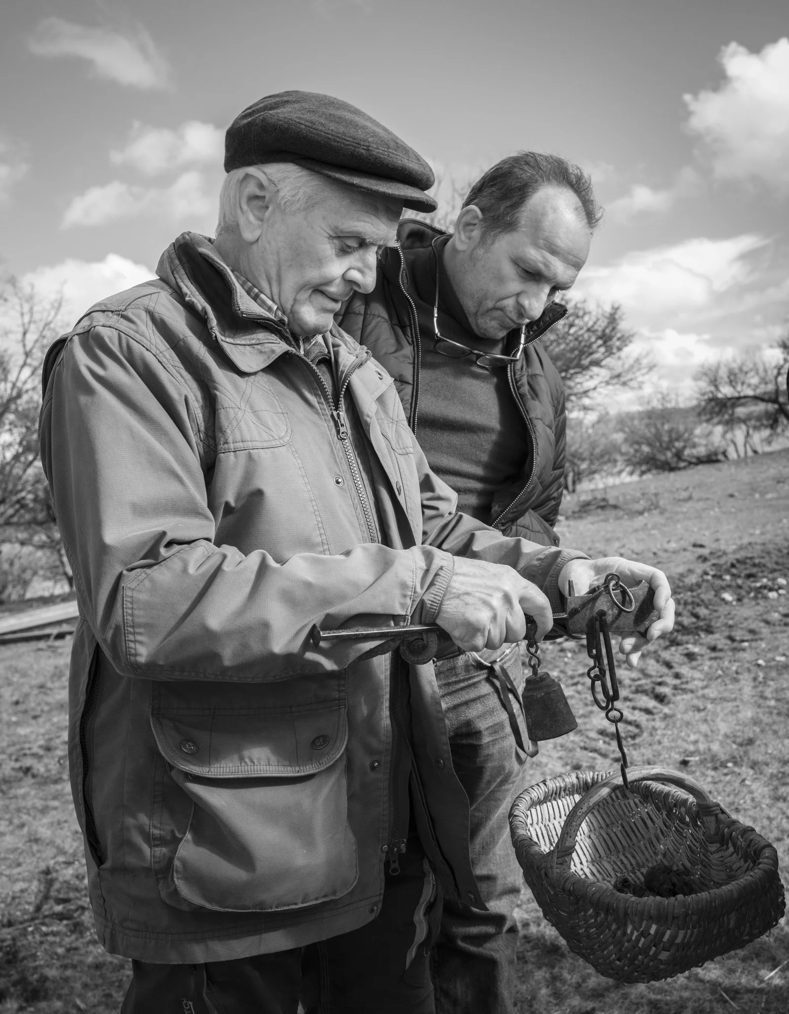 Deux hommes en plein air, l'un d'eux tenant une balance en col de cygne avec un panier en osier suspendu, en noir et blanc.